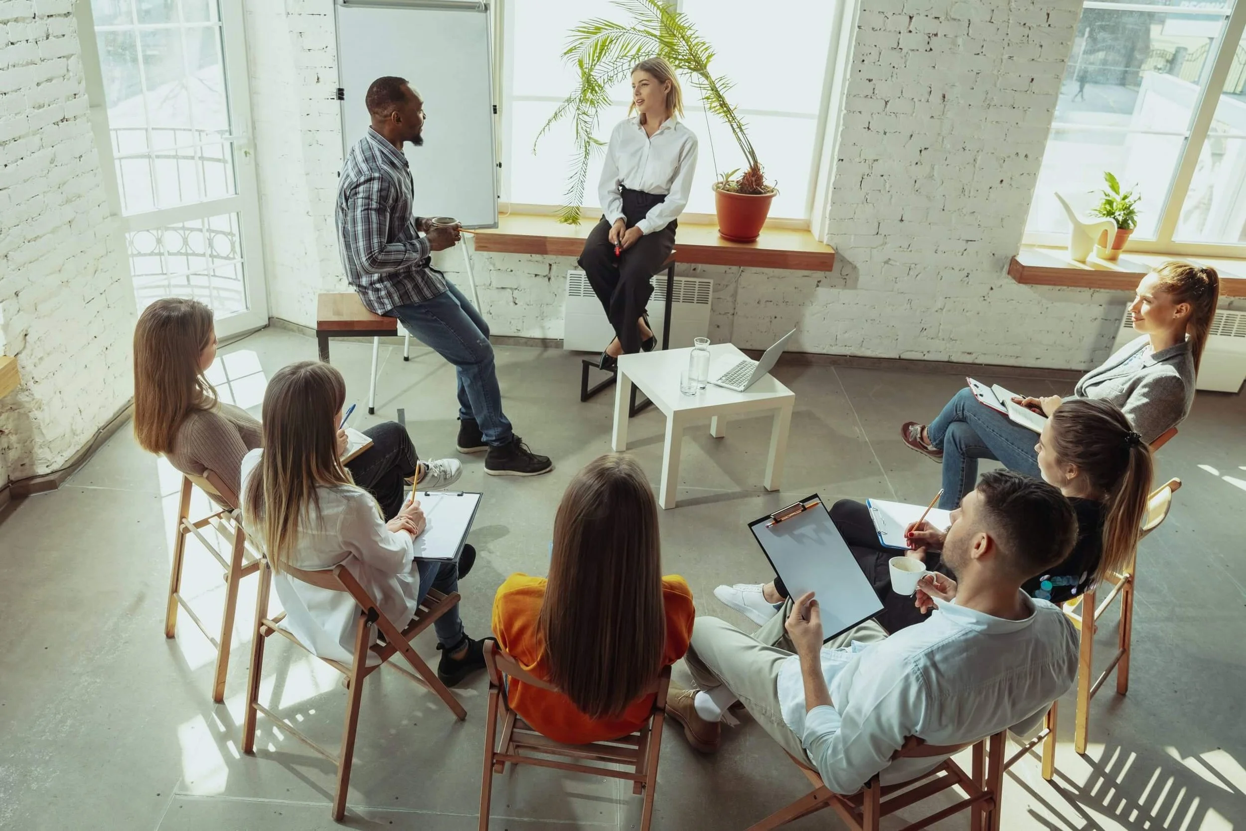 Group of people seated in a circle during a workshop or discussion, with one man speaking and a woman listening while others take notes.