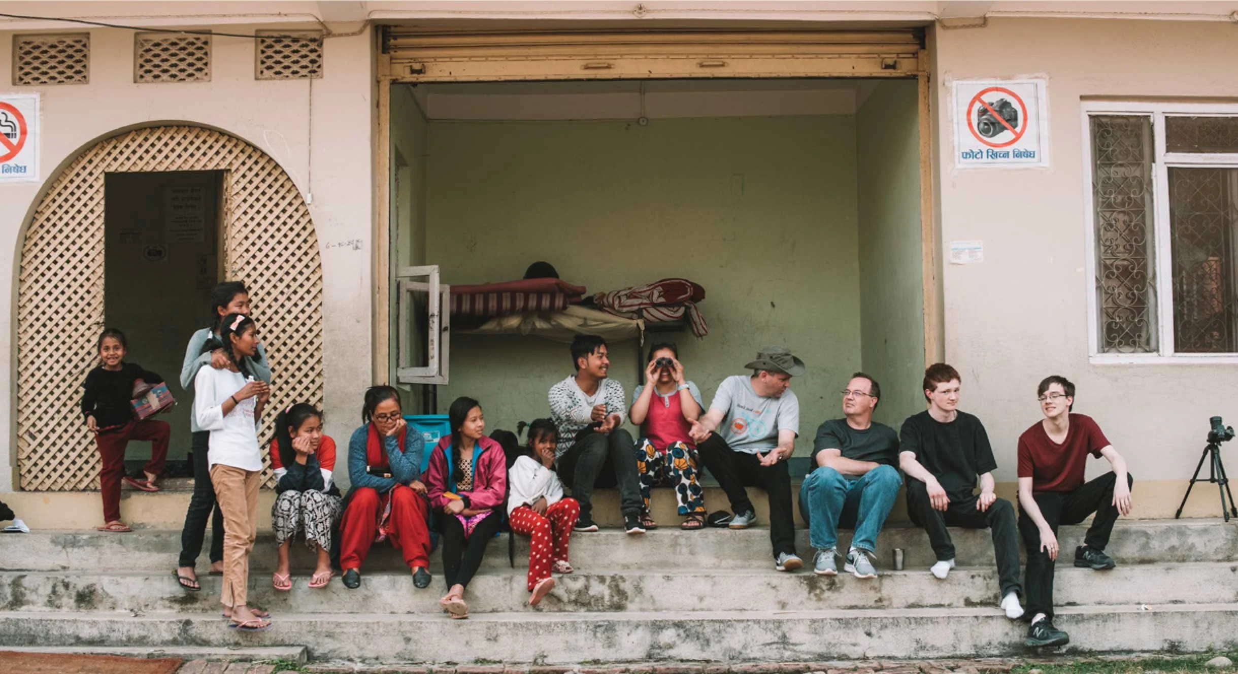 A group of people sitting on steps outside a building, with a no smoking sign and a no camera sign visible on the wall.