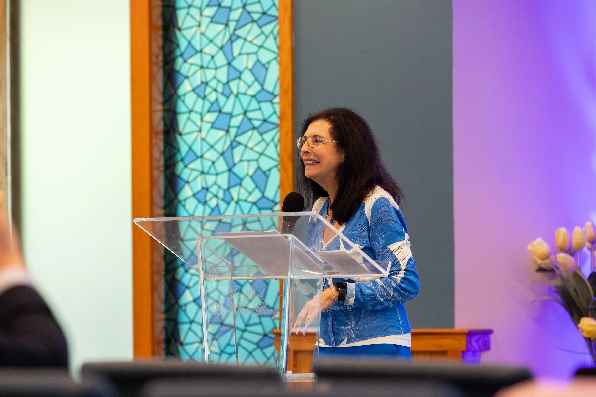 A woman with dark hair and glasses speaking at a clear podium with a microphone in a church or conference setting, with stained glass artwork behind her and lit purple lights to the side.
