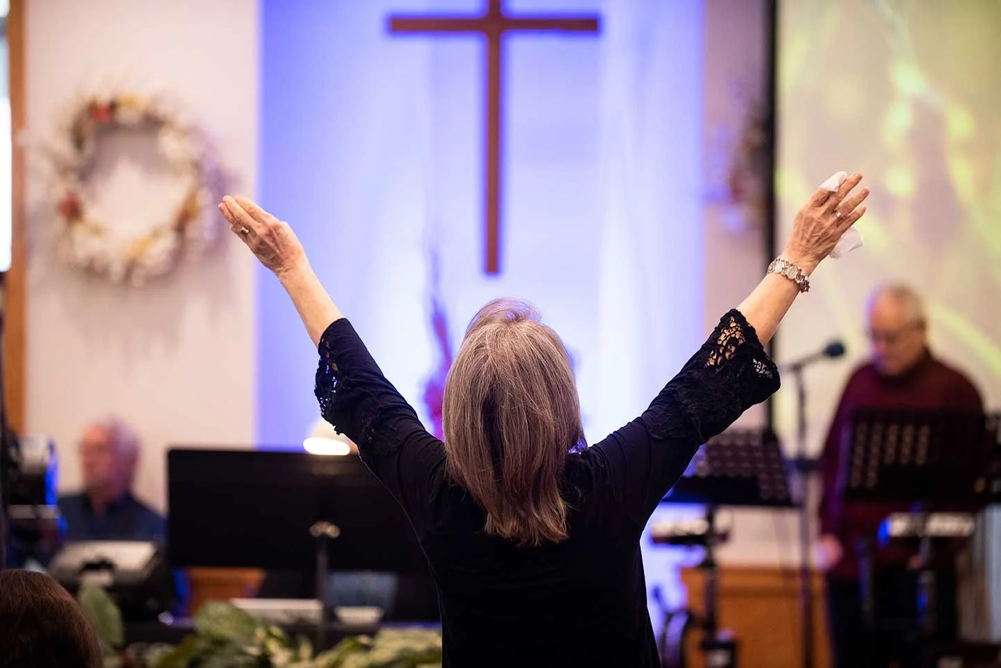 A person with arms raised in a church service, cross on wall in background.