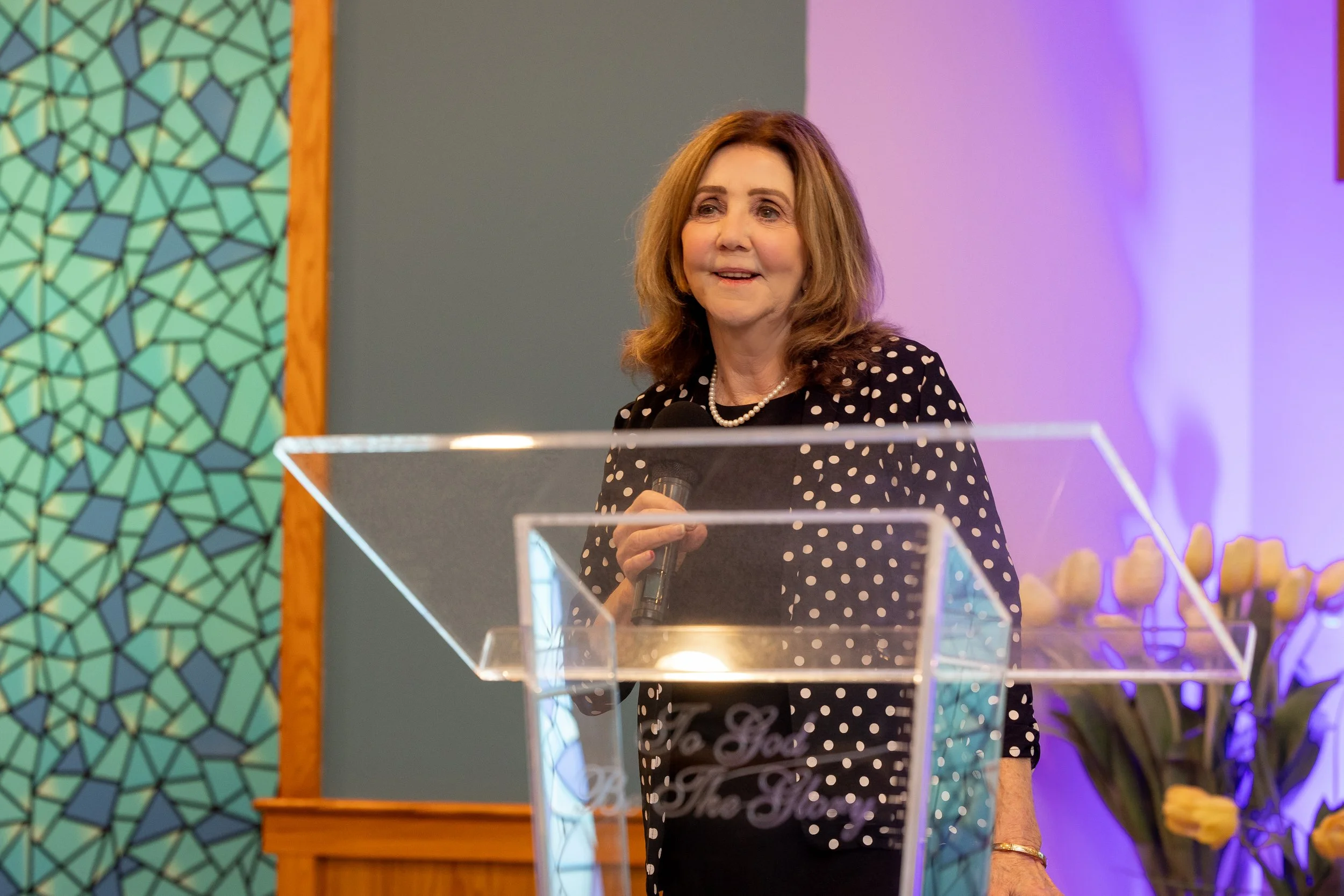 A woman with shoulder-length brown hair, wearing a black polka dot blazer and pearl necklace, speaking into a microphone behind a clear pulpit, in a room decorated with a mosaic wall and yellow tulips.