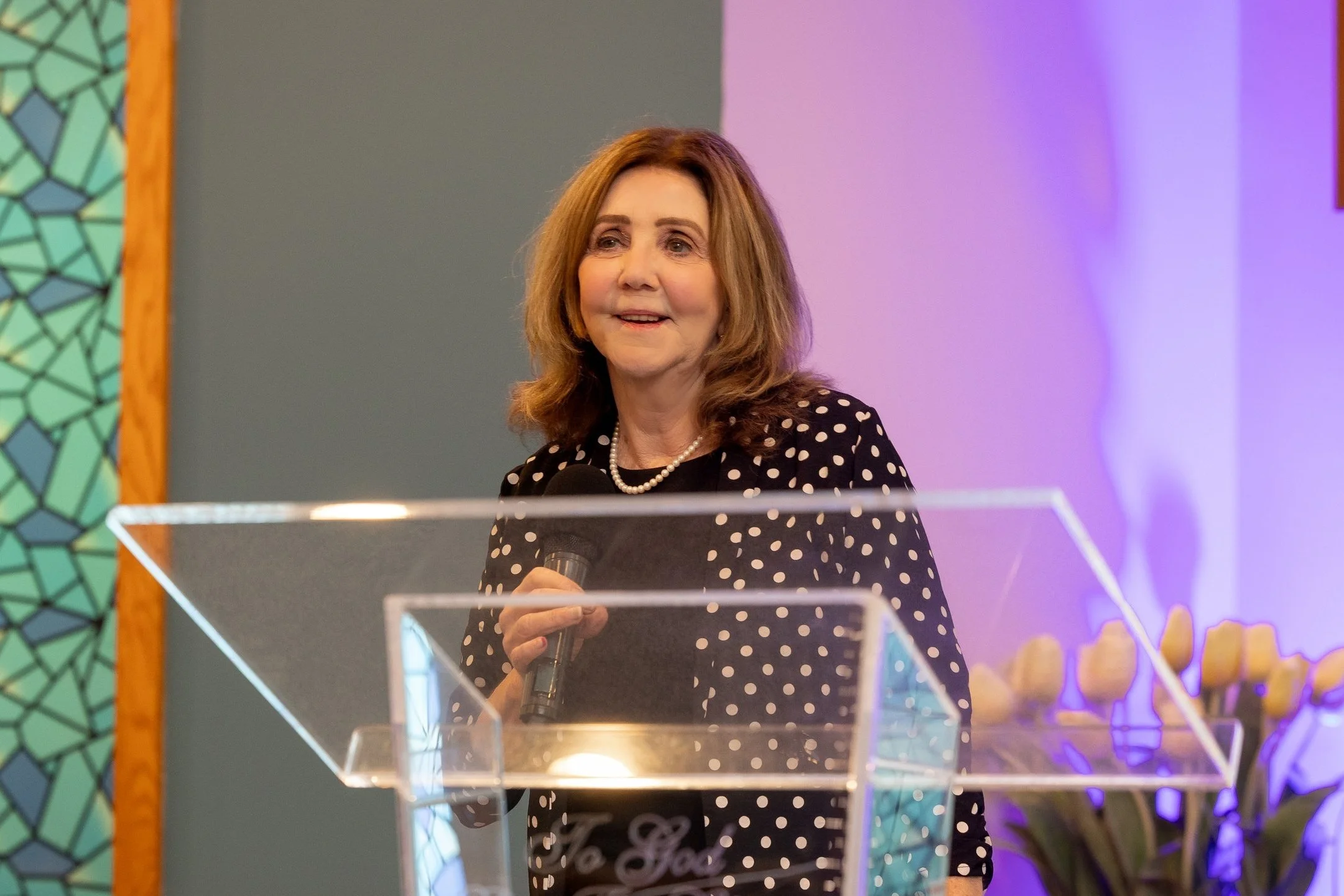 A woman speaking into a microphone during a formal event, with a floral arrangement in the background.