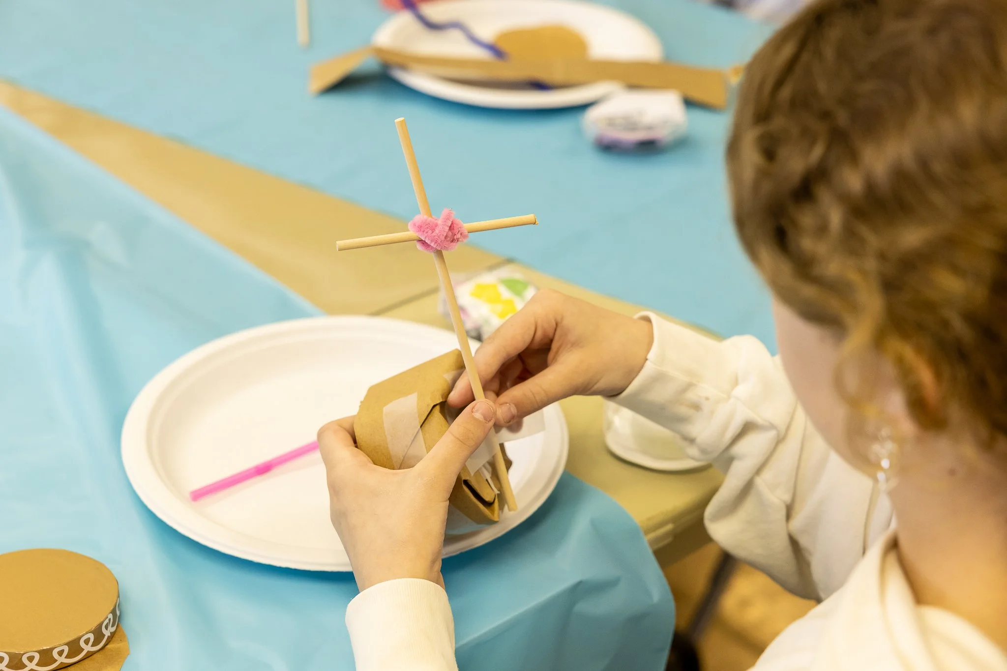 Child making a small craft with brown paper, cross made from sticks, and pink pipe cleaner at a table covered in blue and yellow tablecloths.