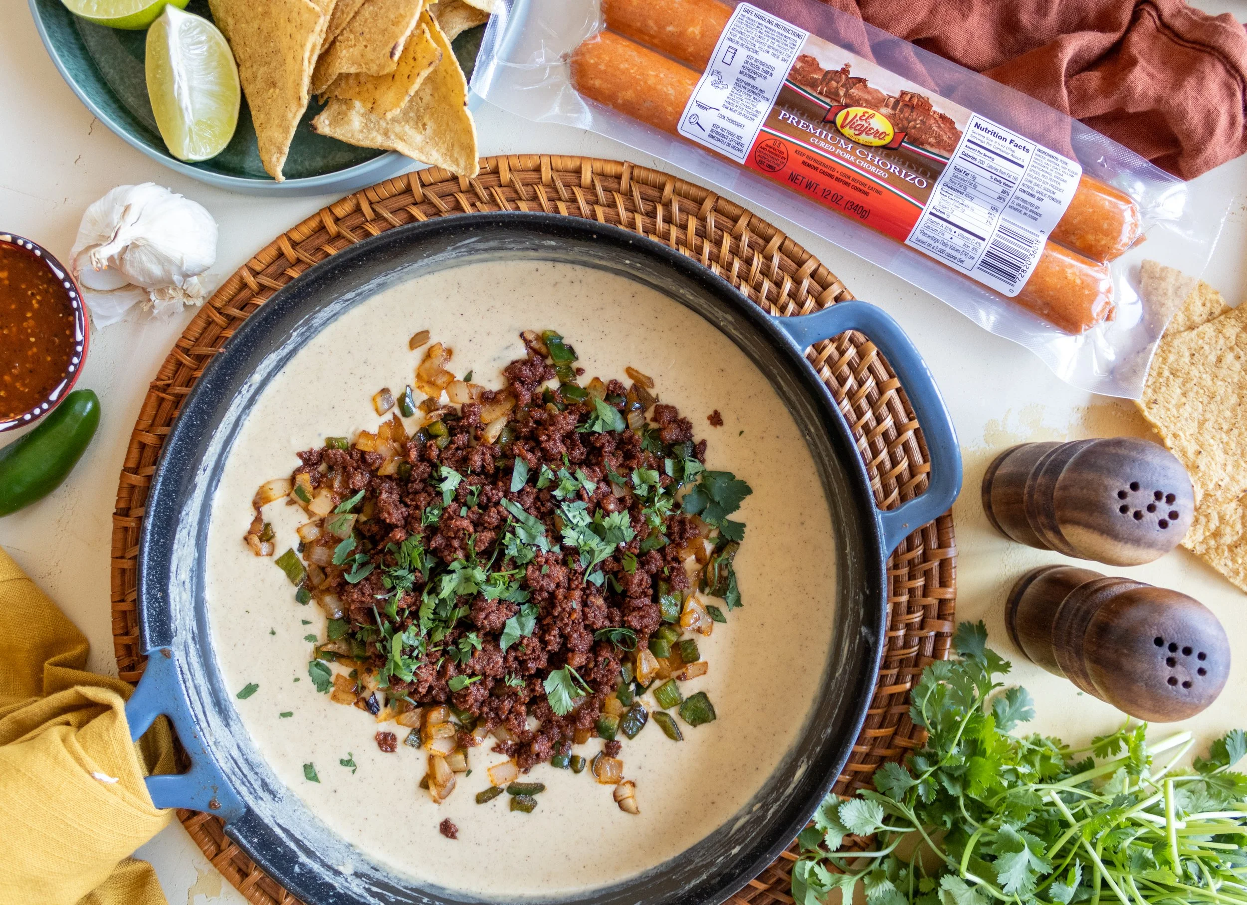 A pot of creamy station with ground beef, onions, and cilantro on top, surrounded by ingredients like limes, avocado jalapeno, garlic, salsa, chips, chorizo sausage, and cilantro, with salt and pepper shakers nearby.