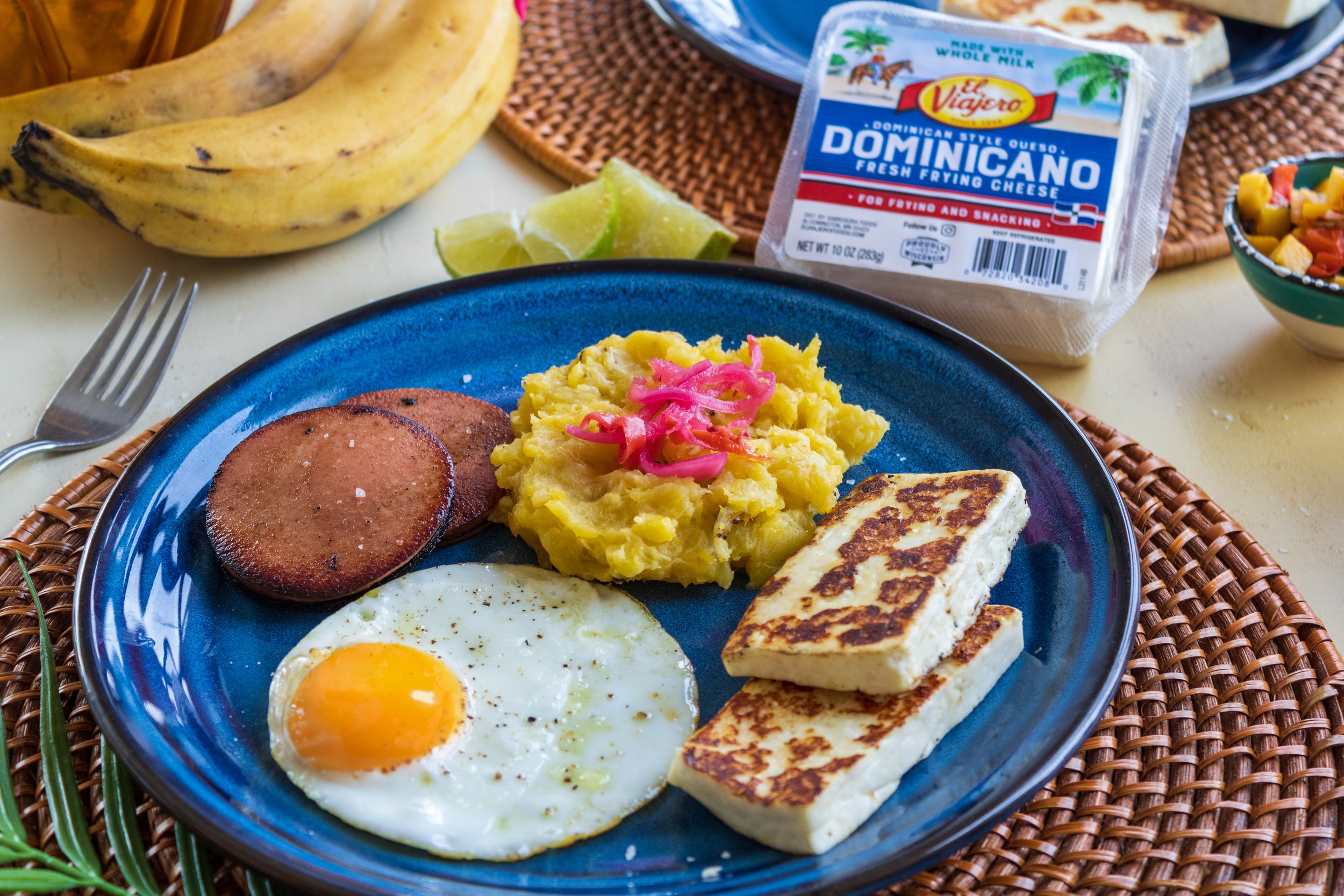 A breakfast plate with a fried egg, sliced sausage, mashed potatoes topped with pink pickled onions, and two pieces of grilled cheese on a blue plate. In the background, there are bananas, lime wedges, a package of Dominican-style fried cheese, and some side dishes.
