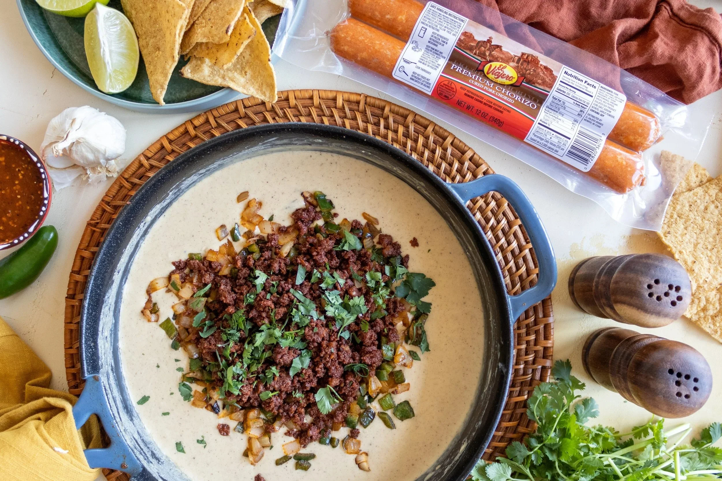 Chorizo, onions, cilantro in a creamy sauce in a black pot. Surrounding ingredients include lime wedges, tacos, garlic, hot sauce, chorizo sausage, tortilla chips, salt and pepper shakers, cilantro, and a garlic bulb.