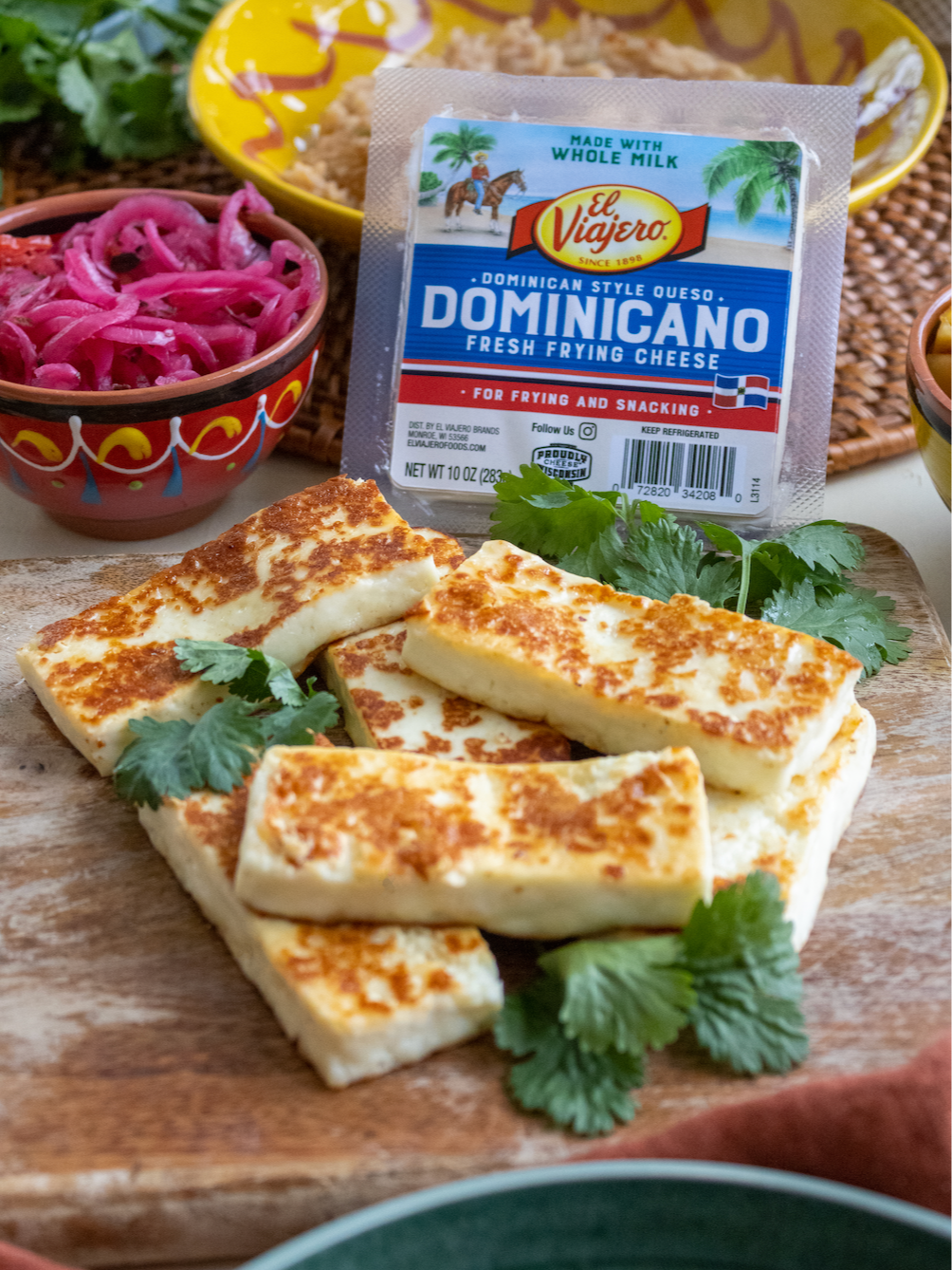 Fried cheese slices garnished with cilantro on a wooden cutting board, with a packet of Dominican-style frying cheese in the background, and bowls of pickled red onions and other toppings nearby.