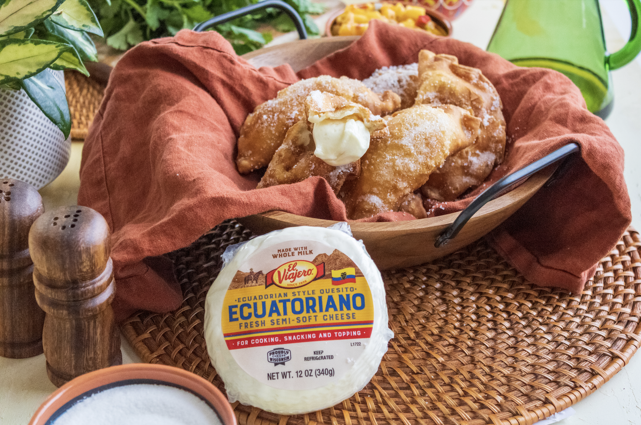 Cheesy empanadas in a wooden basket lined with a rust-colored cloth, with a package of Ecuadorian semi-soft cheese in front, surrounded by spices and green plants.