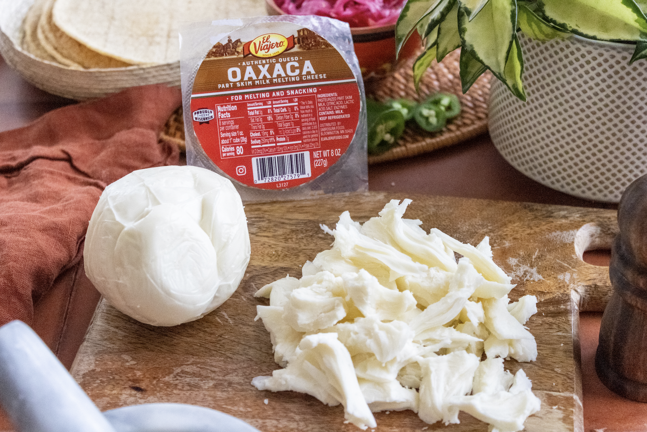 Fresh mozzarella cheese, shredded mozzarella cheese, and a package of shredded Oaxaca cheese on a wooden cutting board, with other kitchen items in the background.