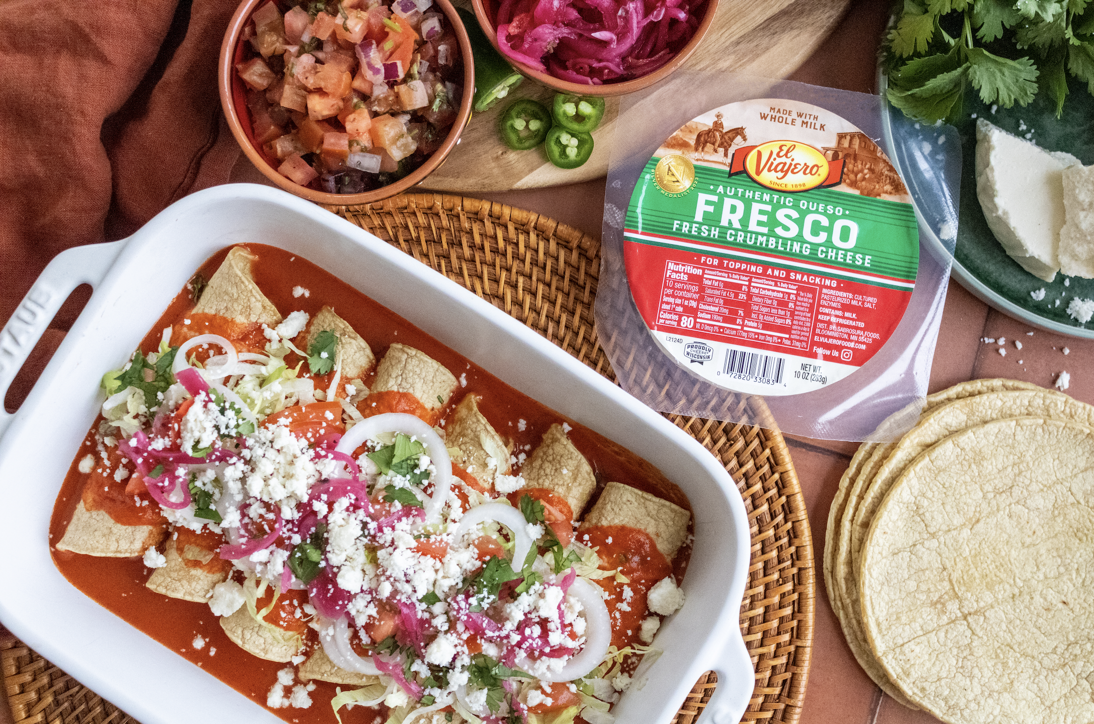 Enchiladas with sliced onions and crumbled Queso Fresco. Surrounding the dish are tacos, a package of Fresco cheese, bowls of chopped tomatoes and onions, sliced jalapeños, tomatoes, and cilantro, with some tortillas on the side.