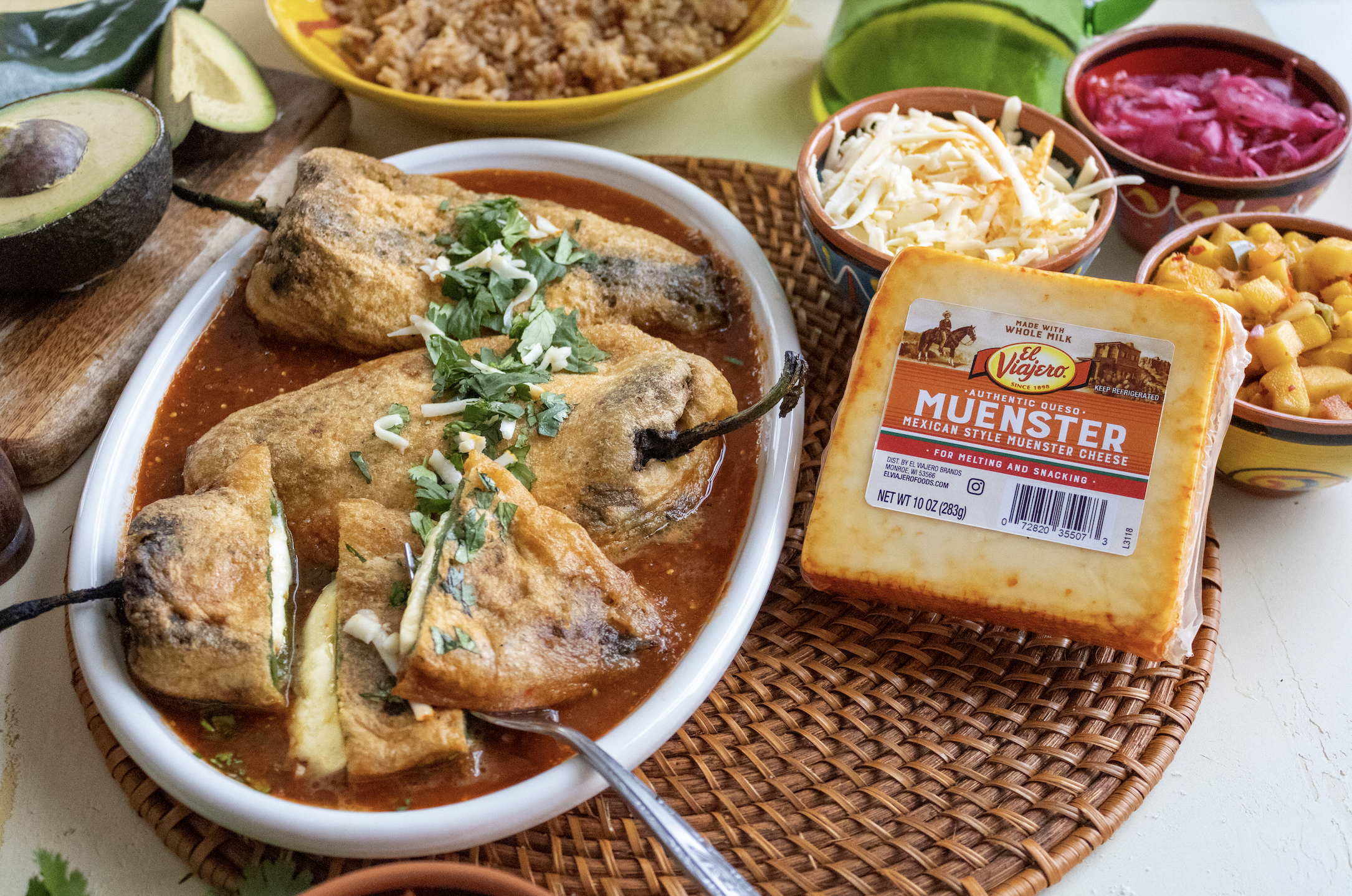 Plate of Mexican stuffed poblano peppers topped with cheese and cilantro, surrounded by bowls of shredded cheese, pickled red onions, corn salad, sliced avocado, and red salsa, with a block of Muenster cheese in the foreground.