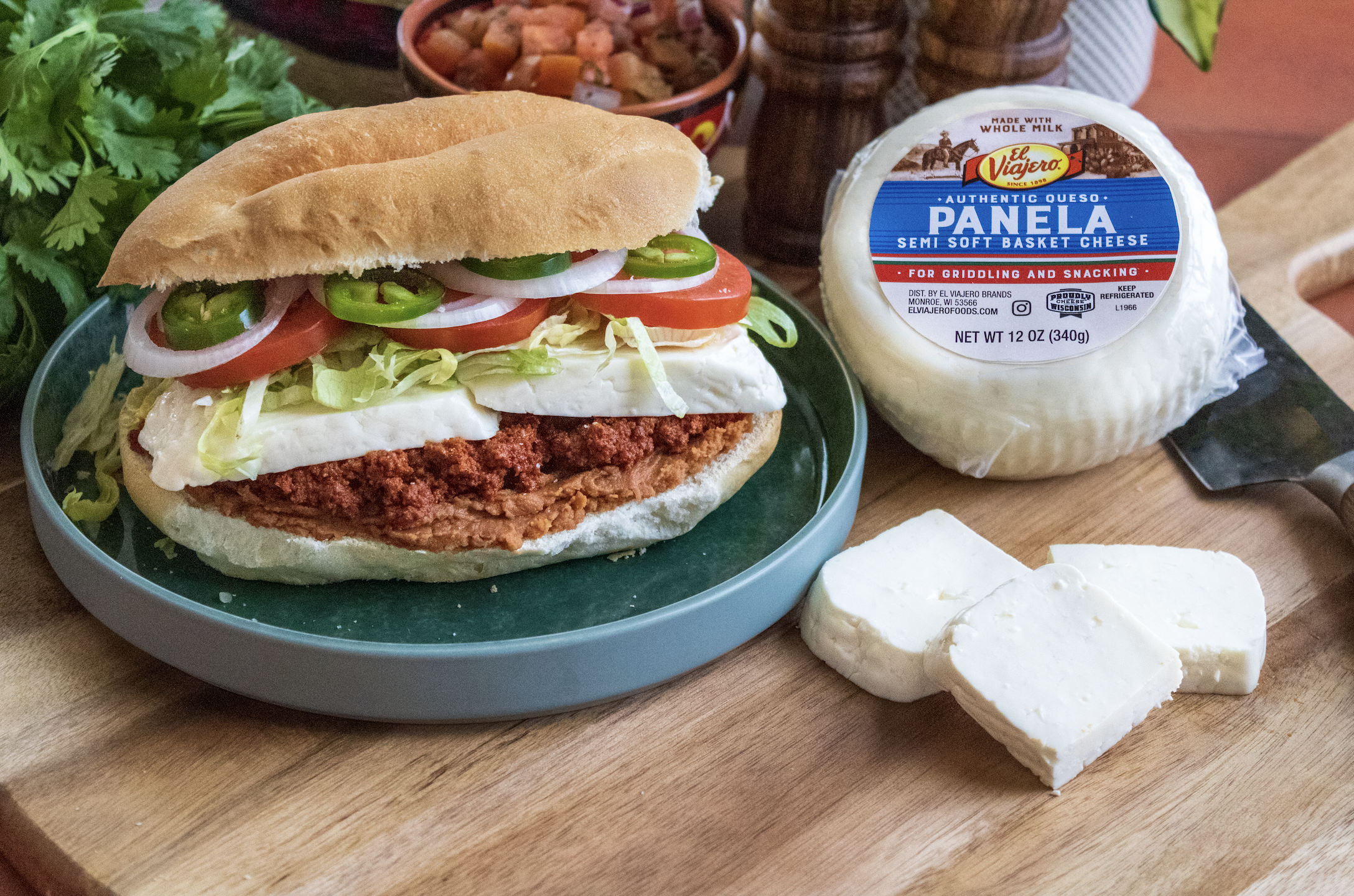 A torta with fried meat, lettuce, tomato, onion, green chili, on a bun, served on a green plate. There is a ball of semi-soft cheese and cheese slices beside the plate. In the background, there are additional condiments and herbs.