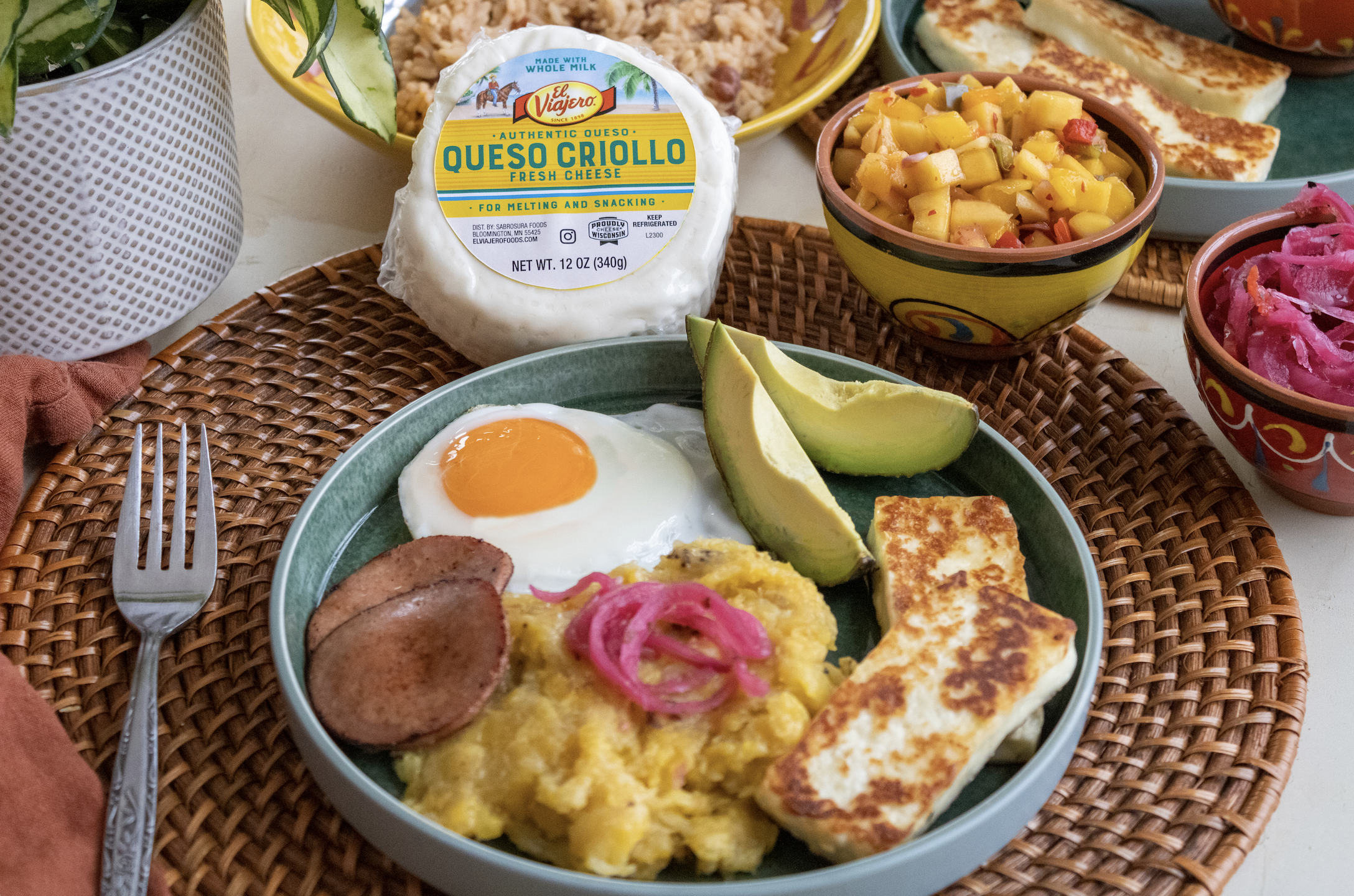 A breakfast plate with a fried egg, slices of sausage, scrambled eggs with pink pickled onions, avocado slices, and fried cheese. Surrounding bowls contain fruit salad and pickled vegetables. A pack of queso criollo cheese is in the background.