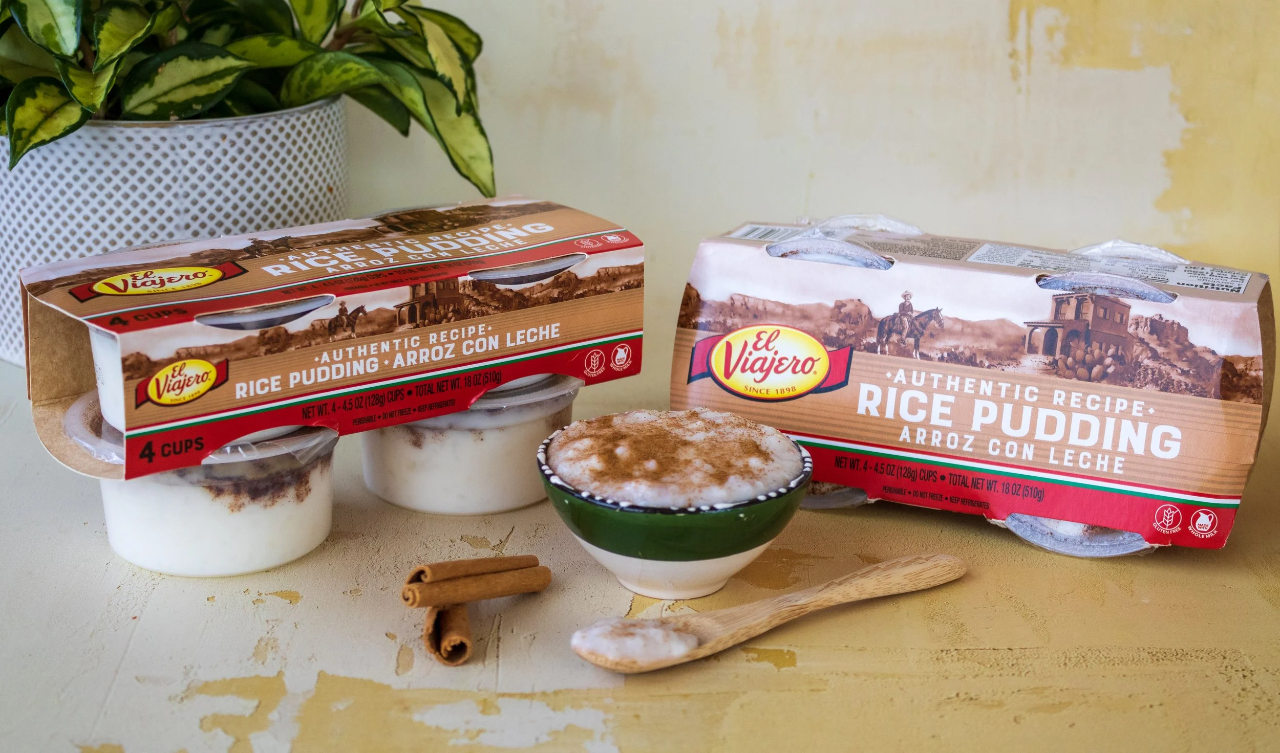 Two packages of El Viajero rice pudding on a table, one opened and one closed, with a bowl of rice pudding topped with cinnamon in front. A wooden spoon and cinnamon sticks are also on the table, and a potted plant is in the background.