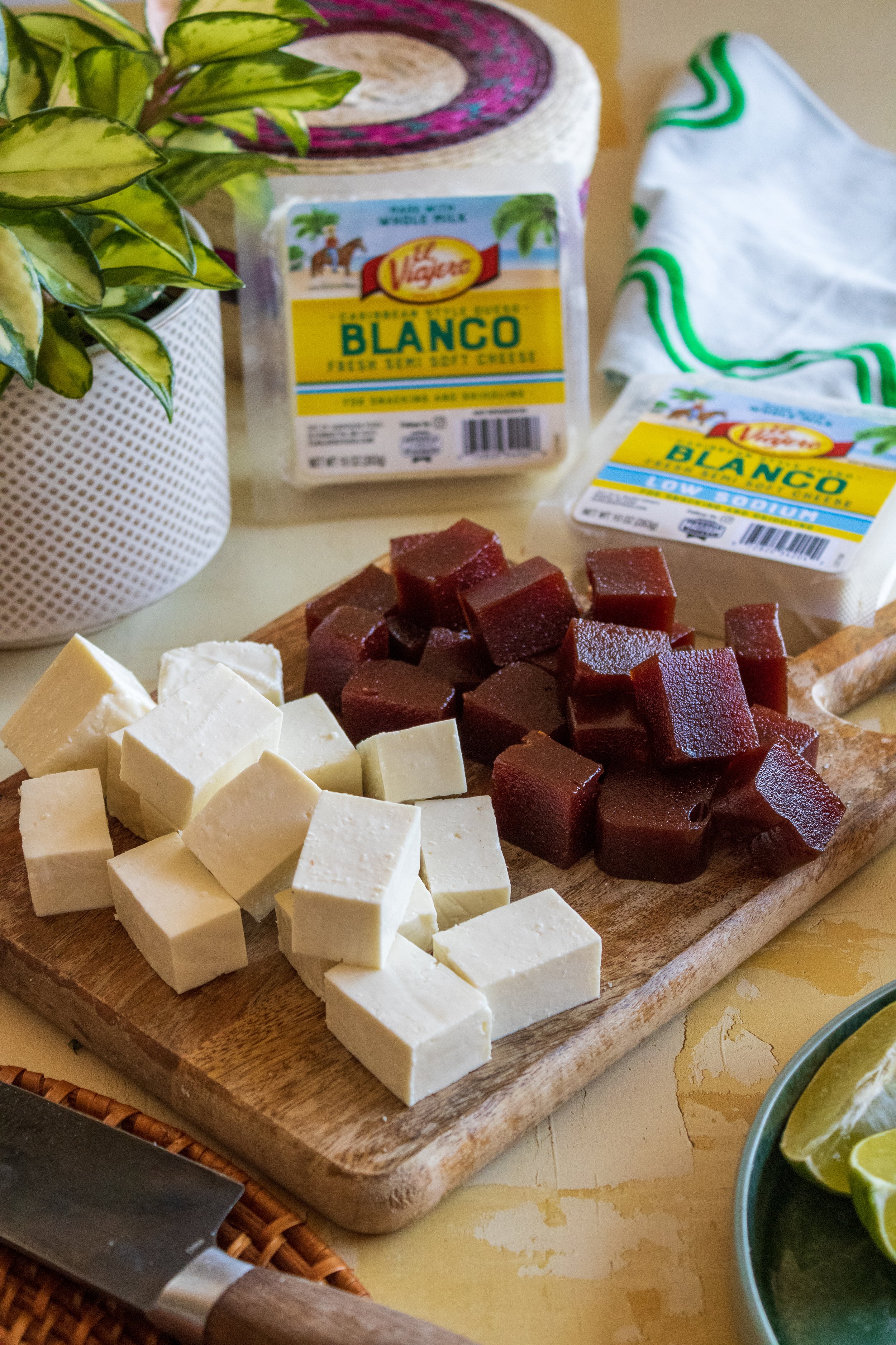 Cubed white cheese and red jellied fruit on a wooden cutting board, with packages of Blanco cheese and a plant in the background.