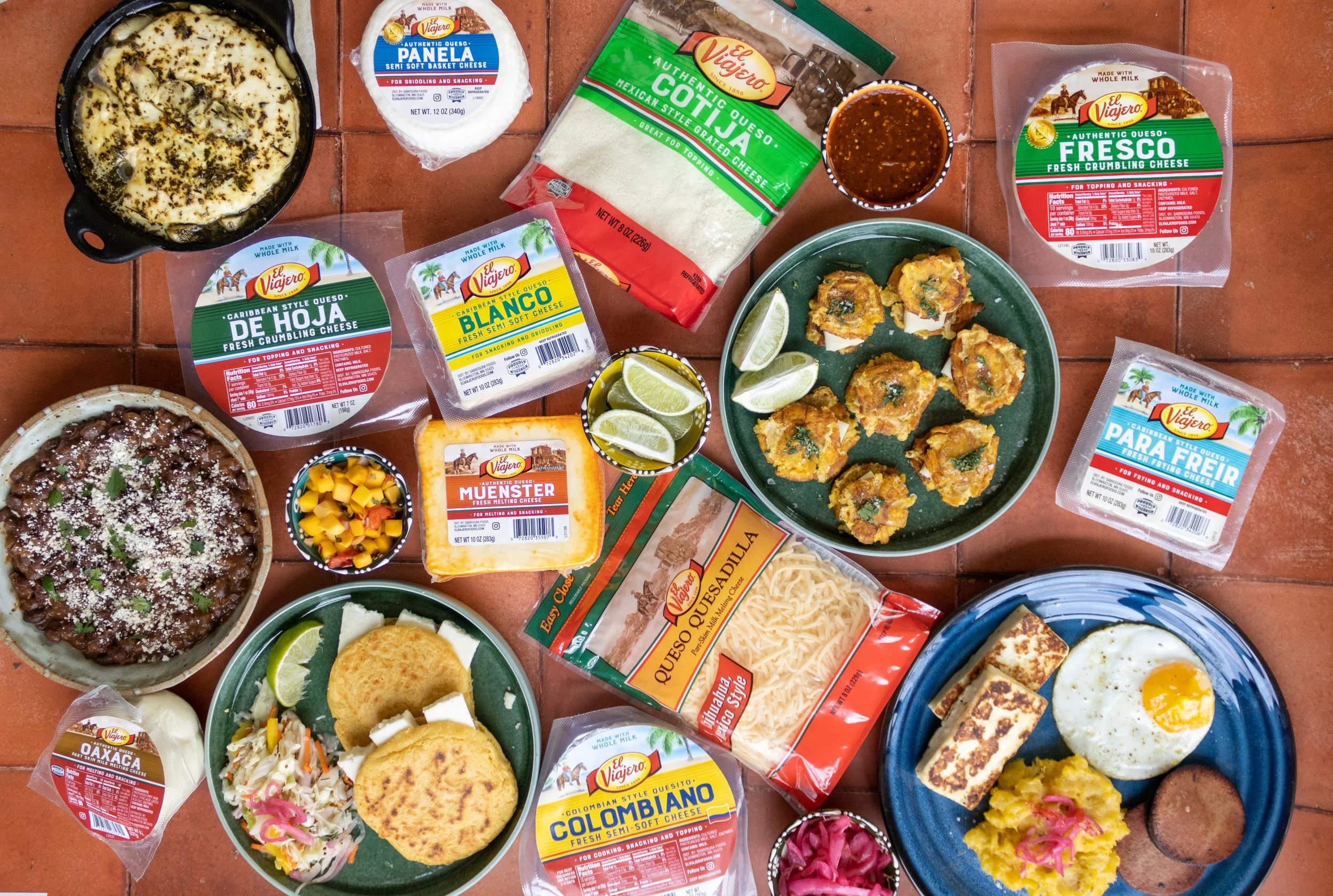 Lay out of El Viajero products and dishes, including quesos, cheeses, mini tacos, fried egg, black bean dish, and lime wedges, all on a terracotta tiled surface.