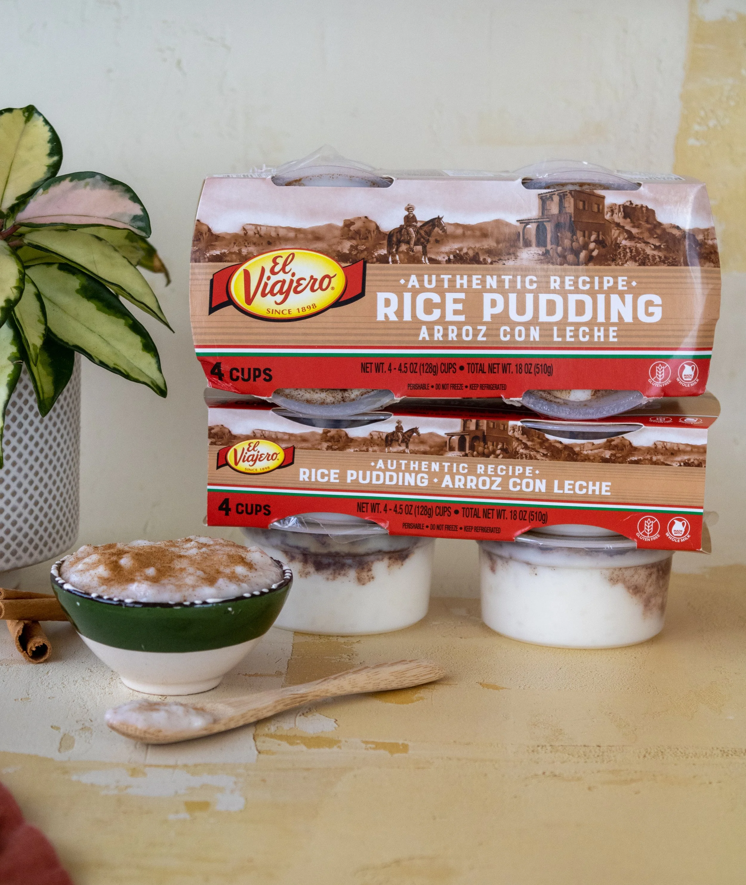 Two boxes of El Viajero rice pudding with cinnamon on top, one bowl of rice pudding with cinnamon and a spoon in front, and a potted plant on the side.