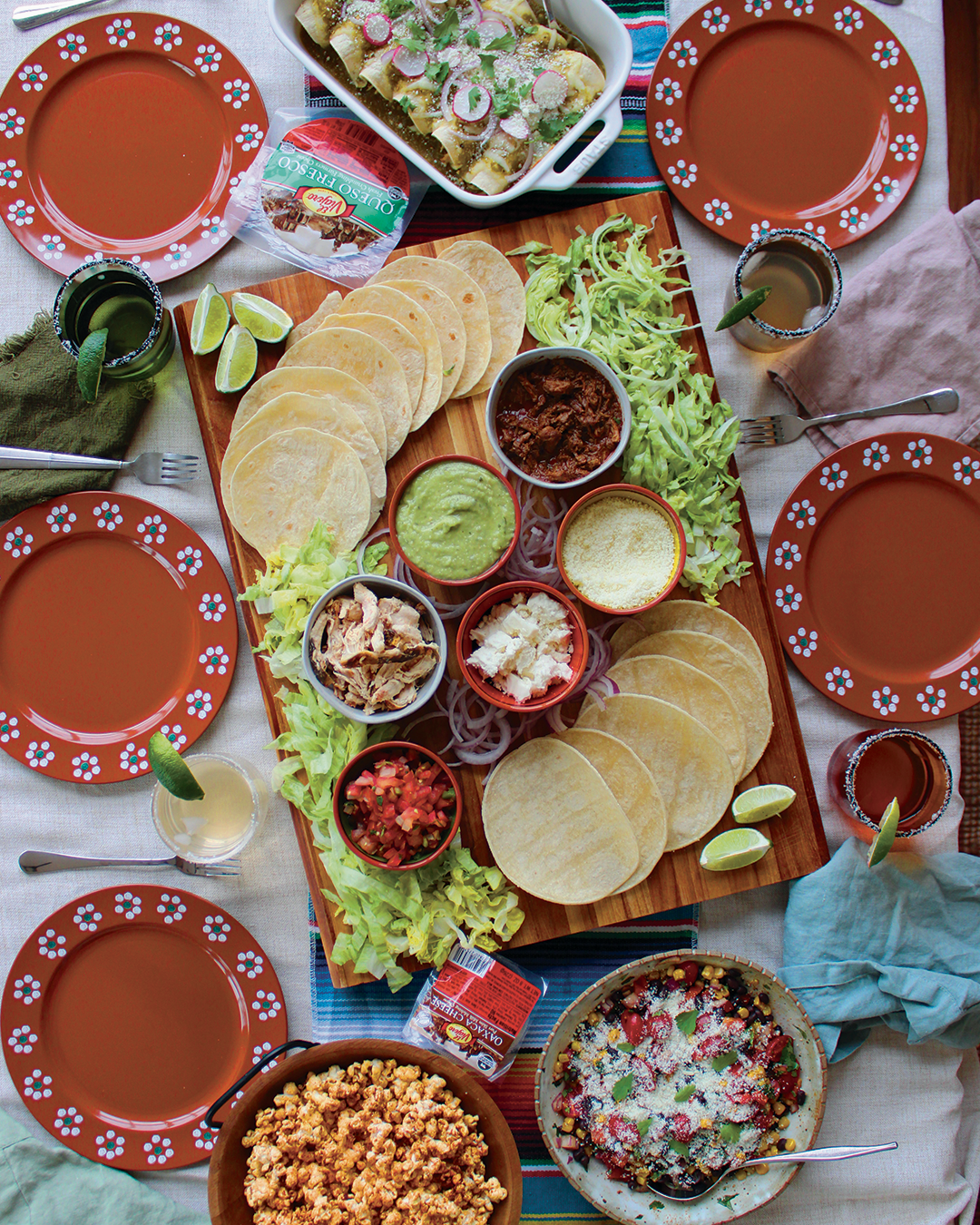 A Mexican feast spread with tacos, tortilla chips, salsa, guacamole, shredded chicken, shredded beef, chopped onions, lime wedges, and a dessert with whipped cream and fruit, along with colorful plates and drinks.