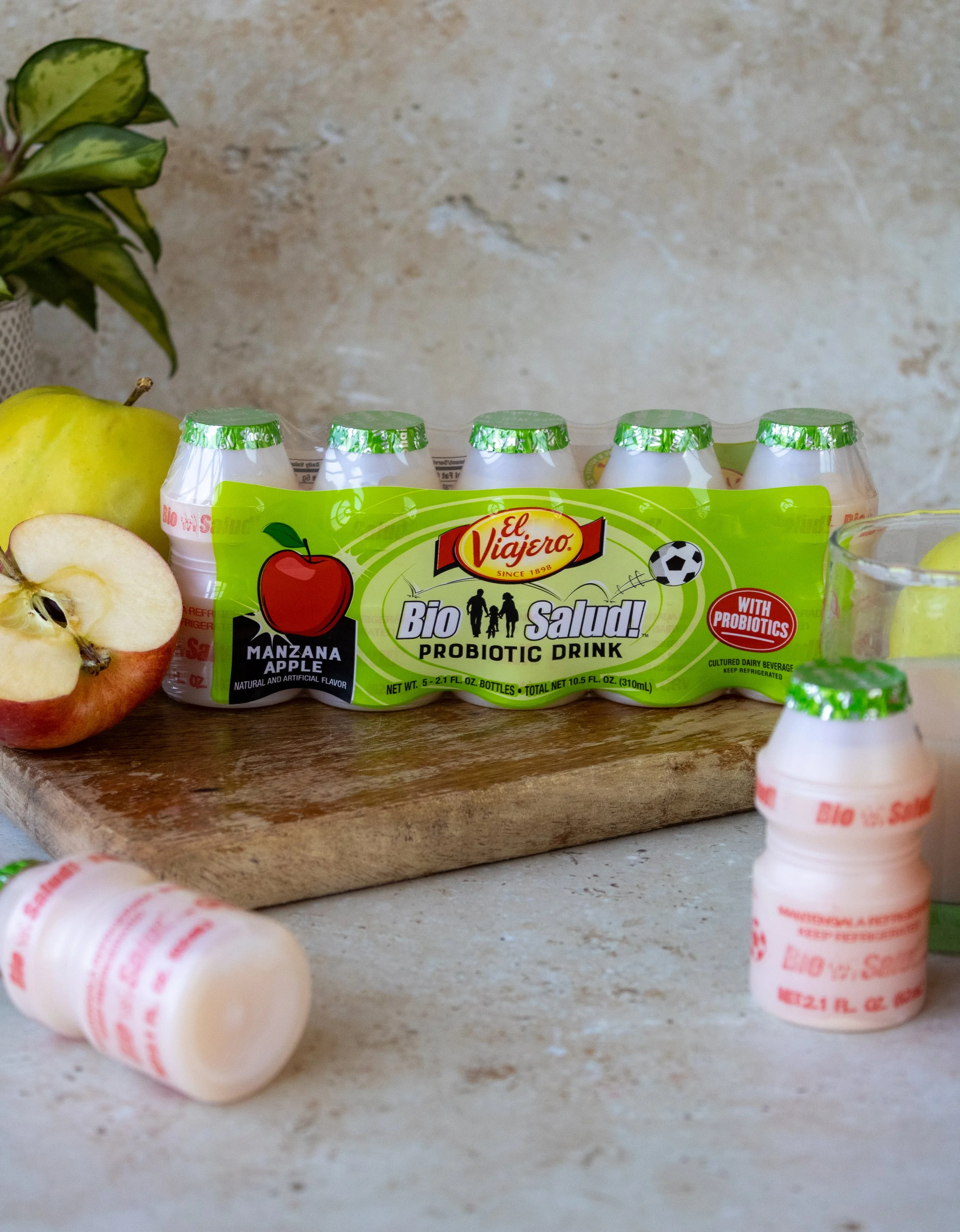 Six bottles of El Viajero Bio Salúd probiotic drink with apple flavor on a wooden surface, with some apple slices and a glass of water nearby.