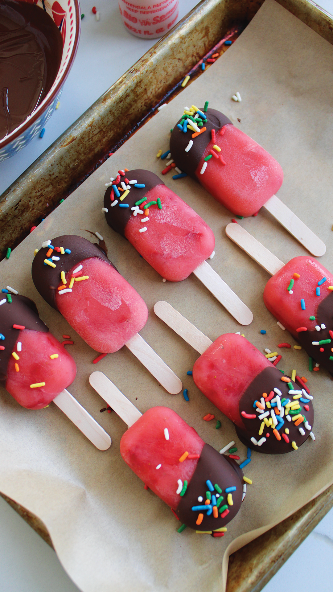 Six pink ice pops with a chocolate-dipped end and colorful sprinkles on top, lying on a baking sheet lined with parchment paper.