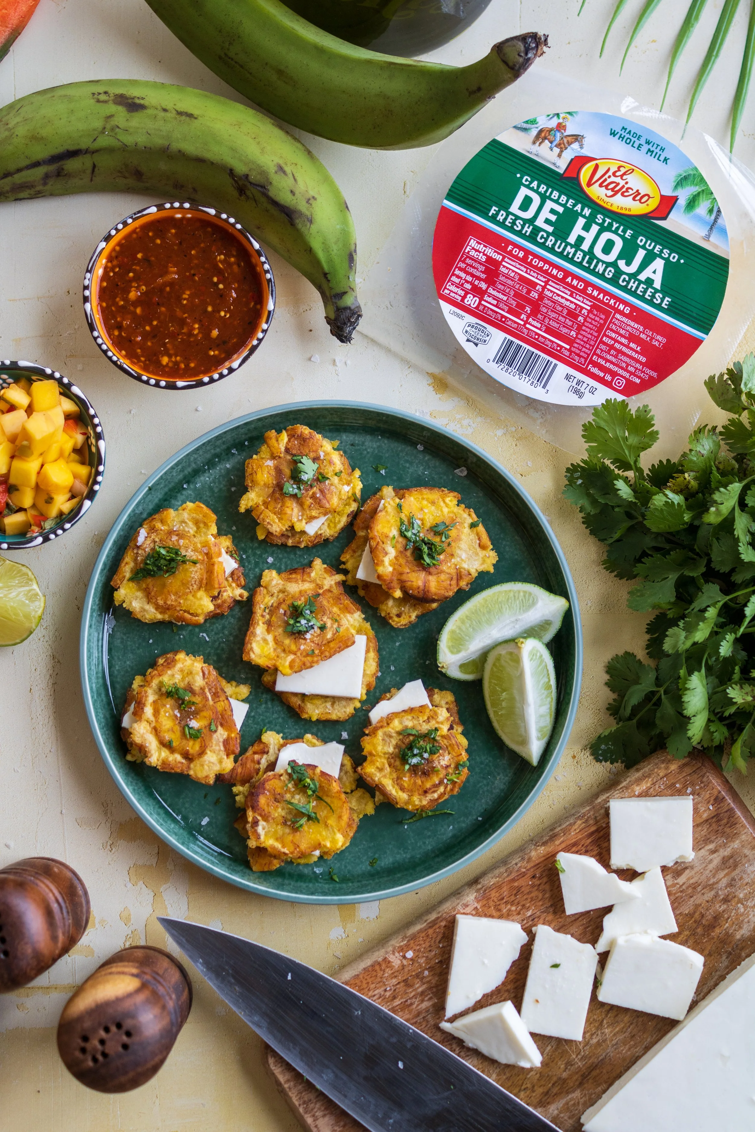 A plate of crispy plantains topped with white cheese and herbs, garnished with lime wedges, surrounded by salsa, cheese, bananas, cilantro, and condiments.