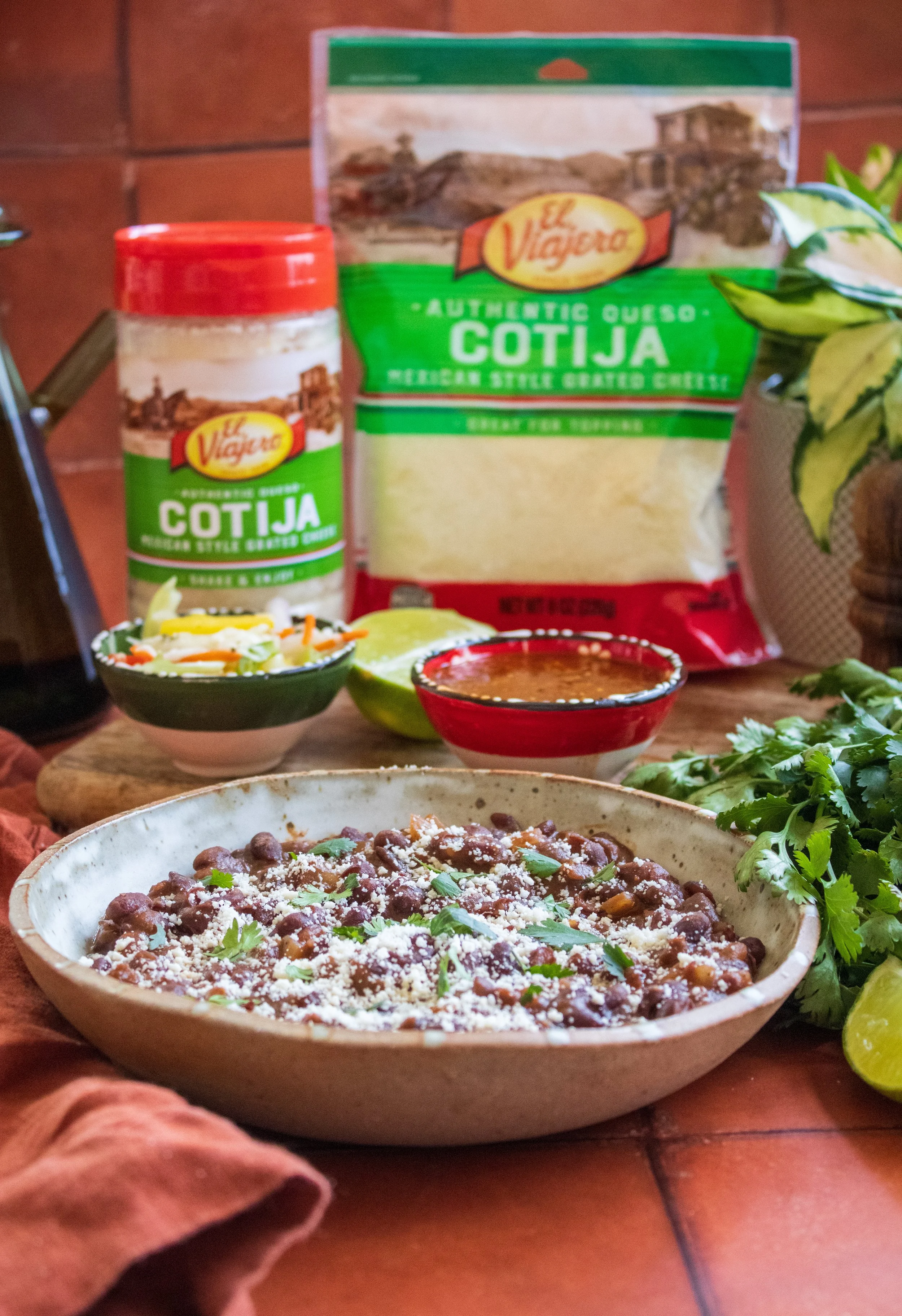 A bowl of black beans garnished with cilantro and cheese, with lime halves, a small bowl of salsa, shredded cheese, and packages of Cotija cheese in the background.