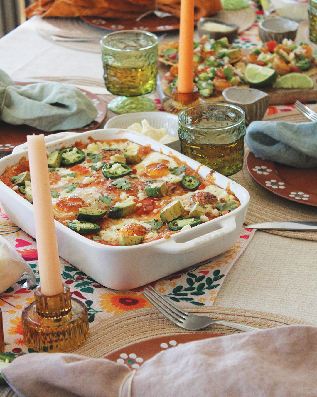 A table set with a baked casserole topped with sliced jalapenos, avocado, cilantro, and cheese, alongside two green drinking glasses, a small bowl of butter, and a serving of salad with lime wedges.