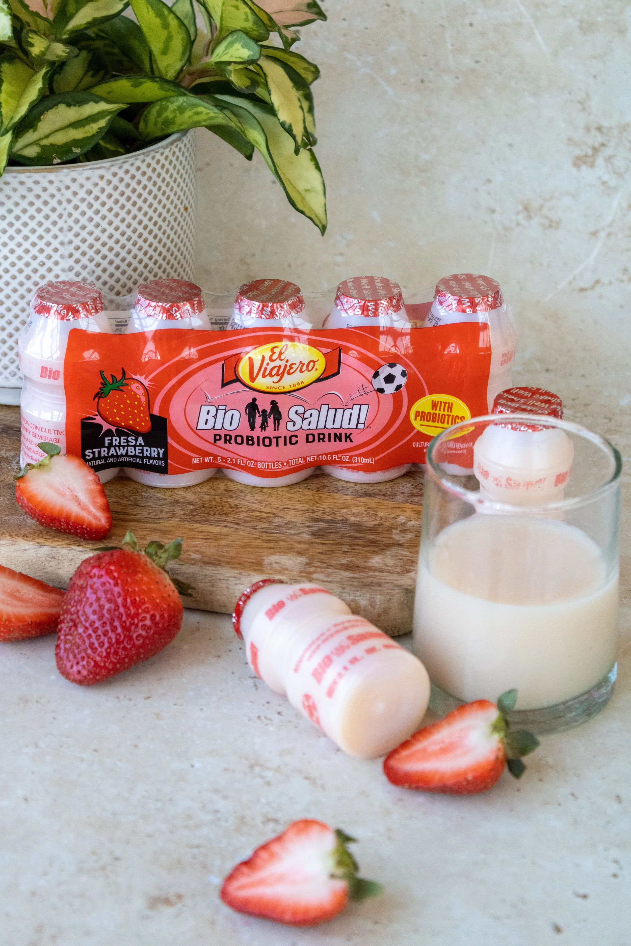 Bottles of probiotic strawberry drink, fresh strawberries, and a glass of milk on a kitchen countertop with a plant in the background.
