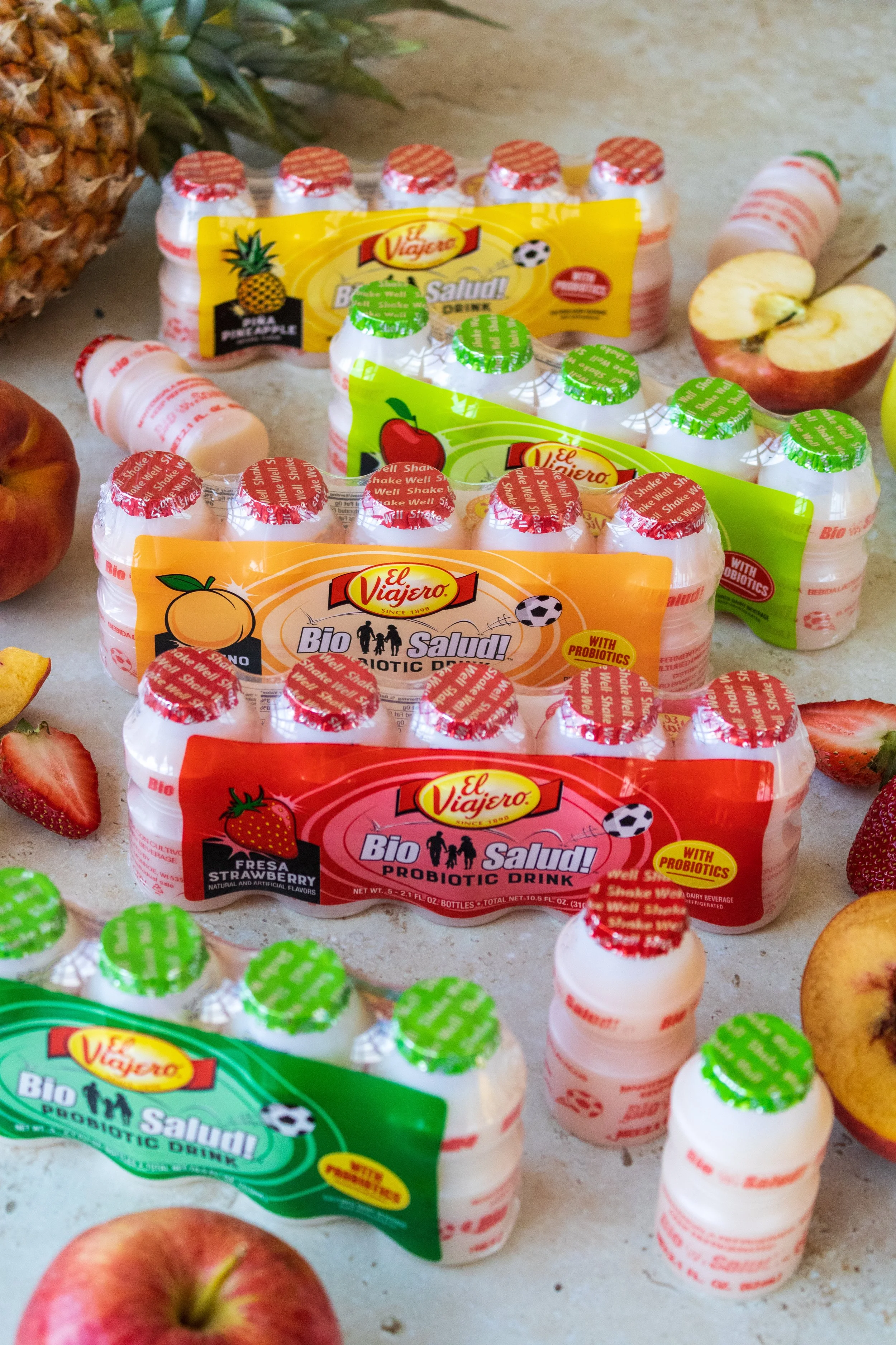 Multiple packages of El Viajero Bio Salud probiotic drinks in various flavors, with apples and strawberries on a light-colored surface.