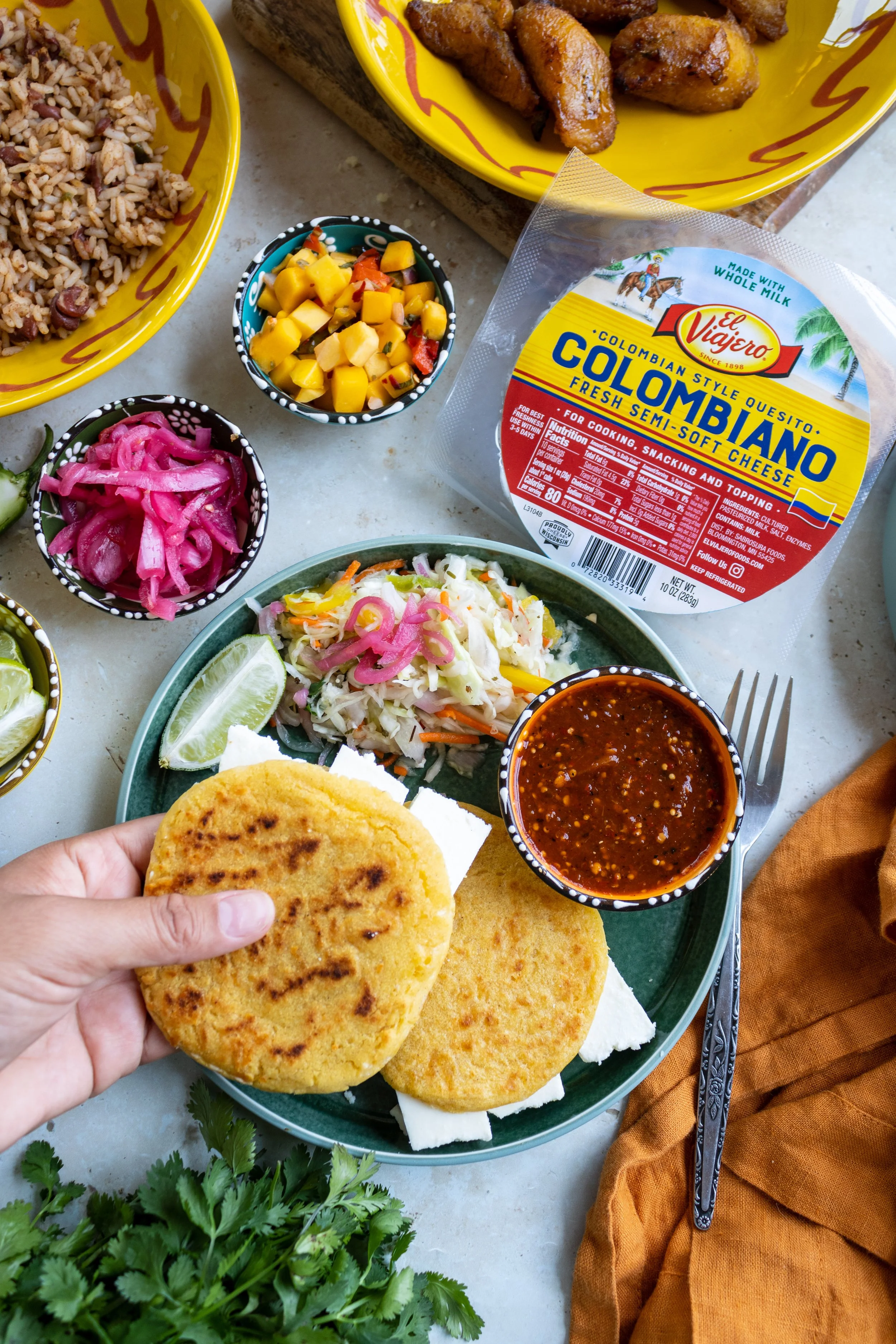 Traditional Colombian meal with fried plantains, rice and beans, pickled red onions, a plate of arepas with cheese, a small bowl of salsa, fresh cilantro, and a package of Colombian-style queso campesino cheese.