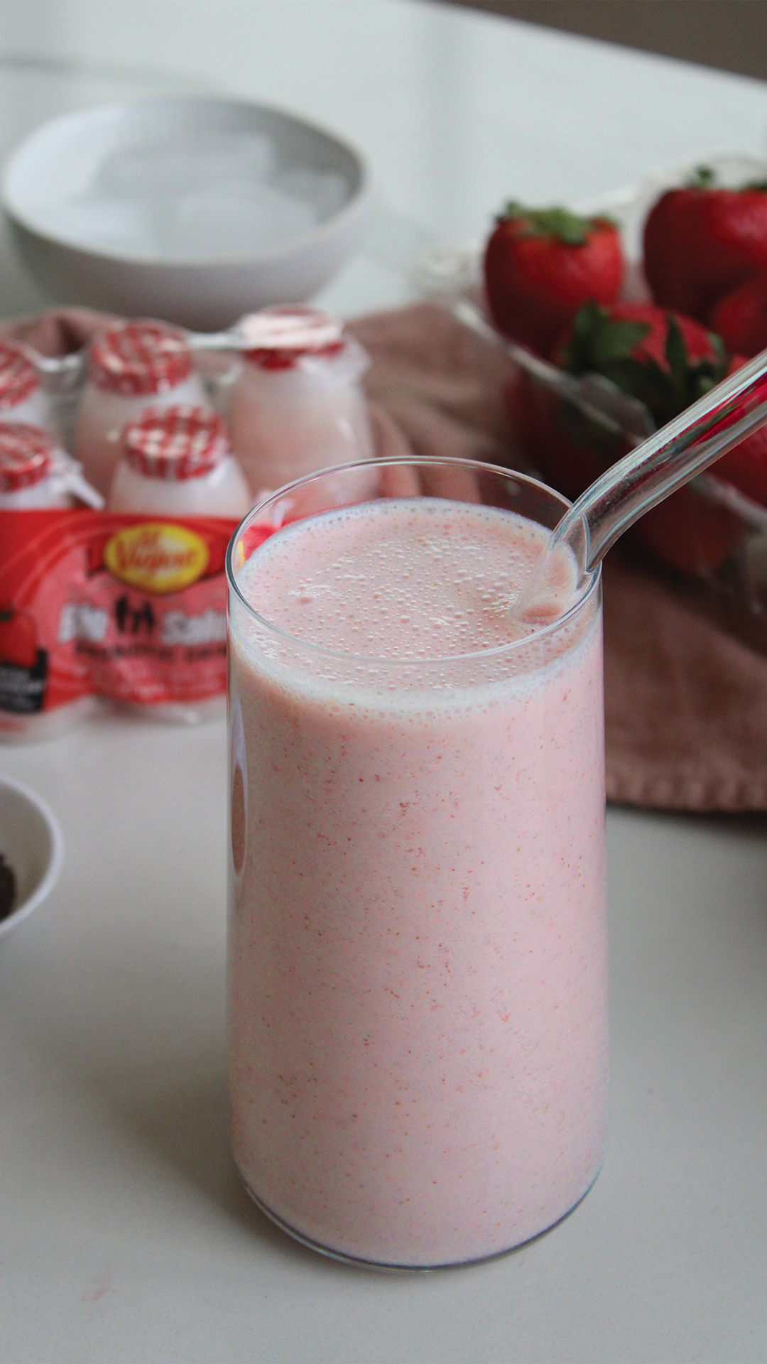 A tall glass of pink strawberry smoothie with a straw, placed on a white surface.