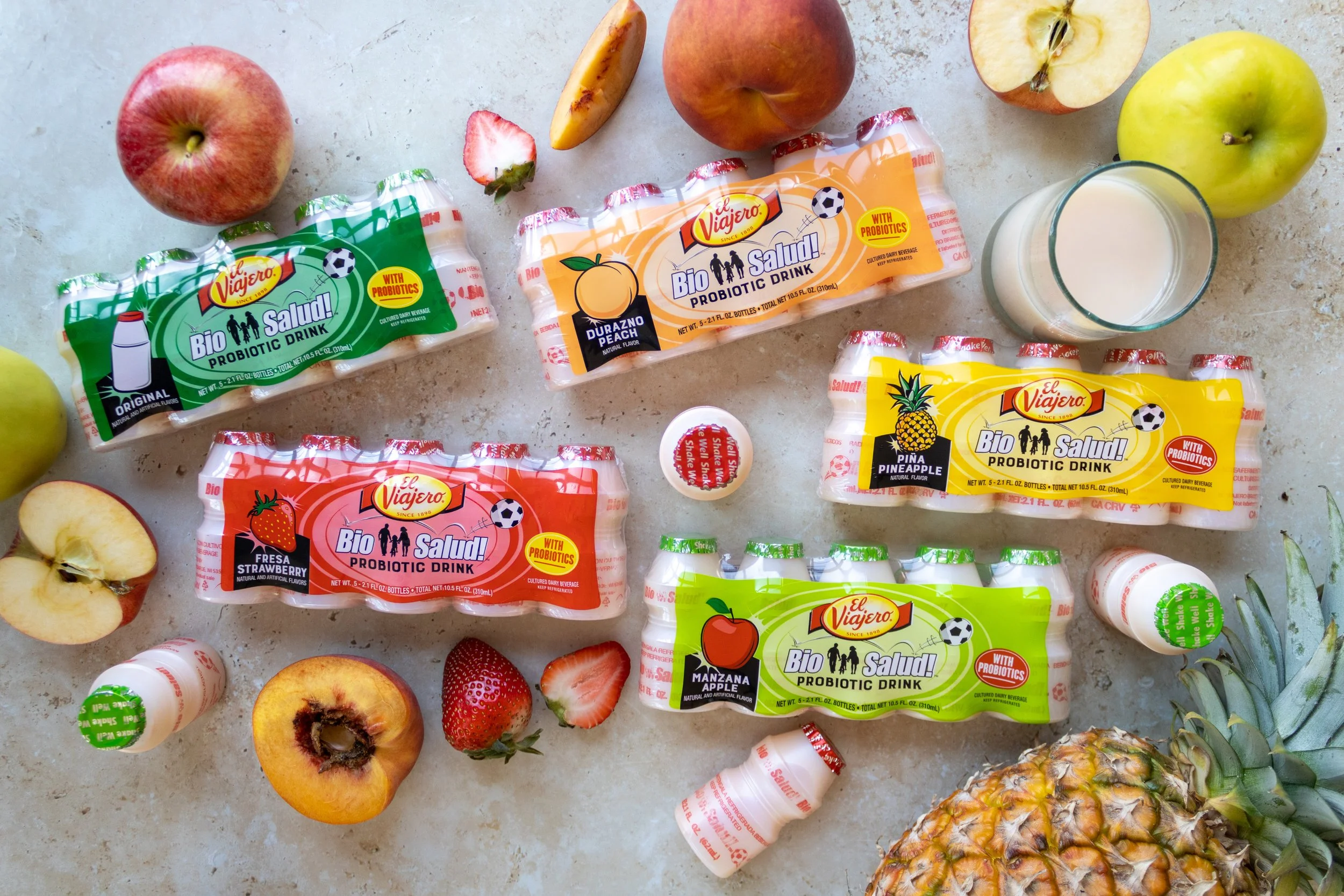 Assorted packages of El Viajero probiotic drinks in various flavors, including strawberry, banana, pineapple, apple, and peach, along with fresh fruits like apples, strawberries, and pineapple, a glass of milk, and small bottles of probiotic capsules on a marble surface.