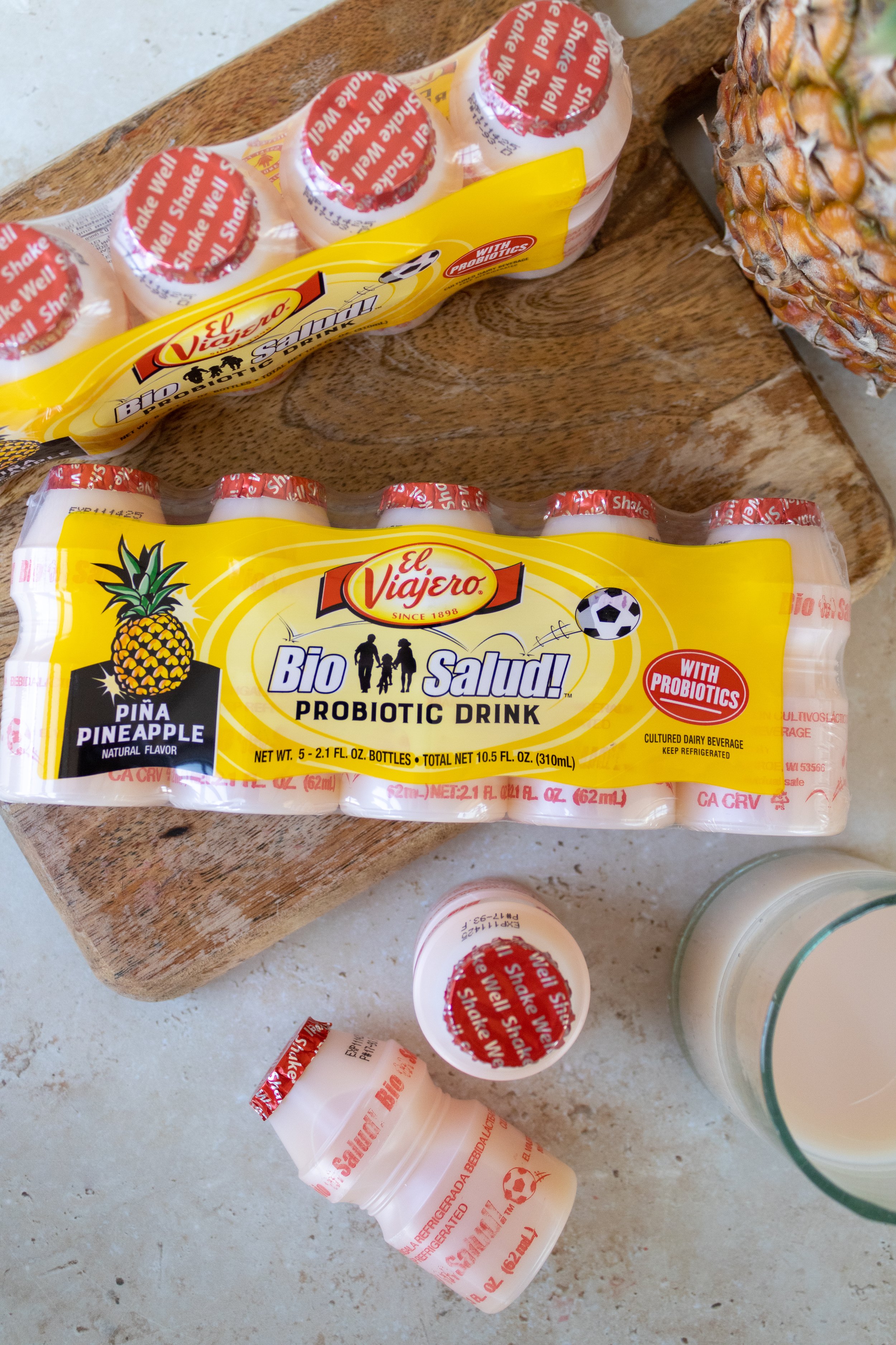 Two packs of El Viajero bio probiotic drink bottles, pineapple flavor, placed on a wooden surface with part of a pineapple and a glass of milk nearby.