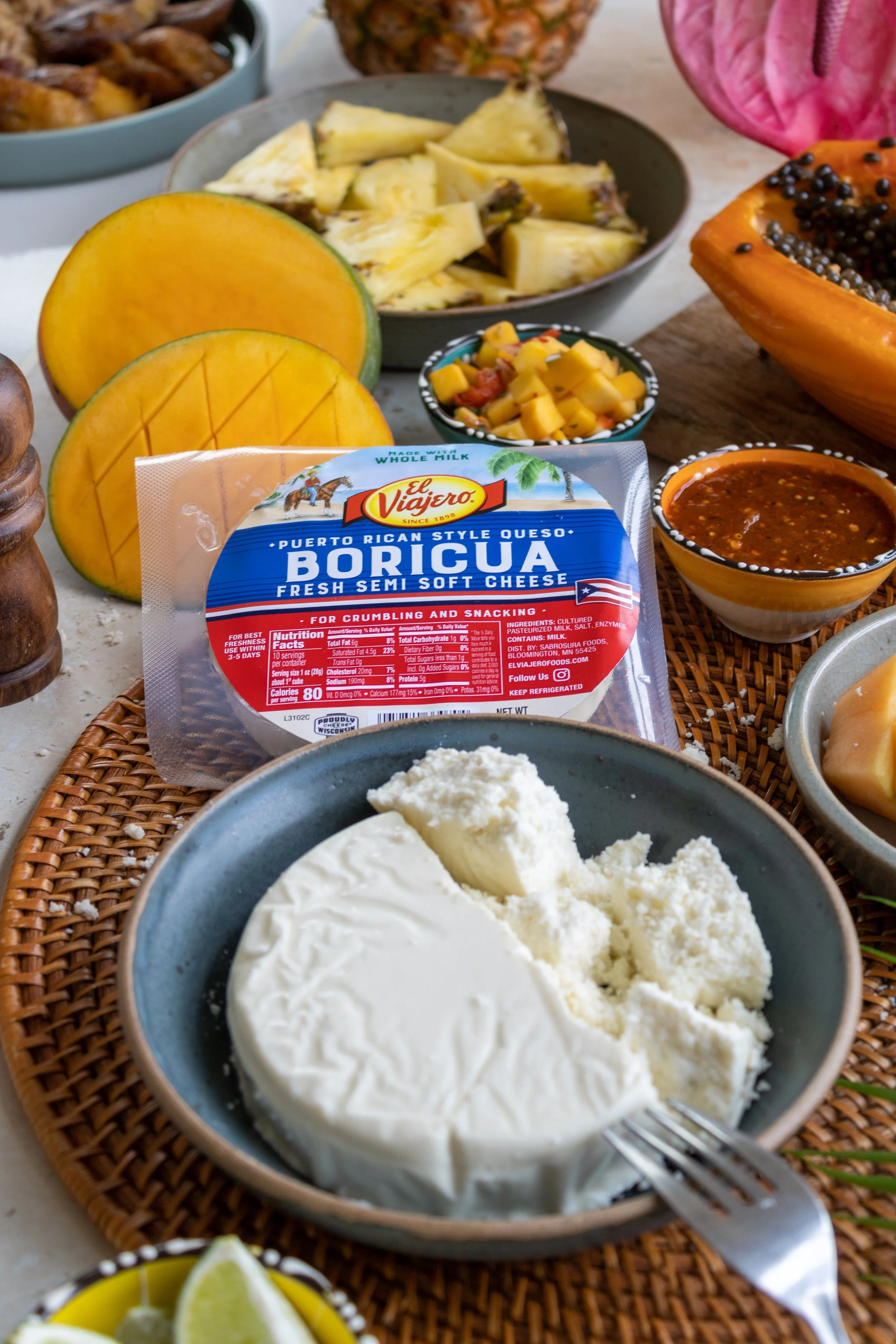An assortment of tropical fruits and cheeses on a table, including sliced cantaloupe, pineapple, cubed mango, and various cheeses like queso fresco and semi-soft cheese. There are also bowls of salsa, black beans, and chopped fruit, along with a package of Puerto Rican style cheese labeled 'Boricua'.