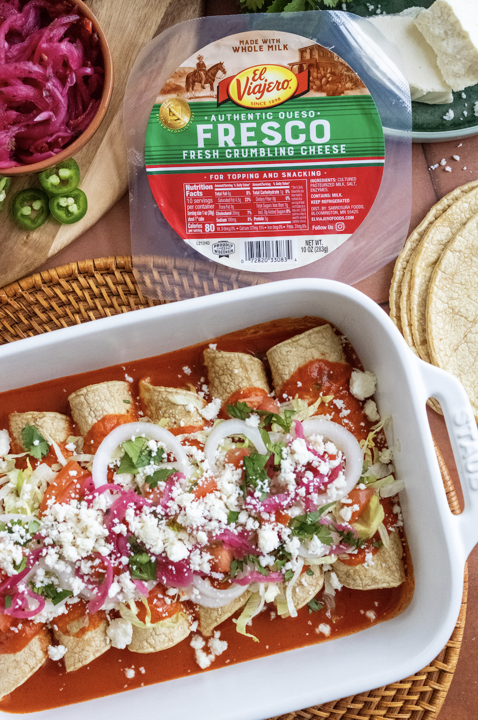Dish of tacos topped with chopped onions, cilantro, crumbled cheese, and sliced tomatoes, in a white serving dish, with a background of pickled red onions, jalapeño slices, tortillas, and a package of crumbling cheese.