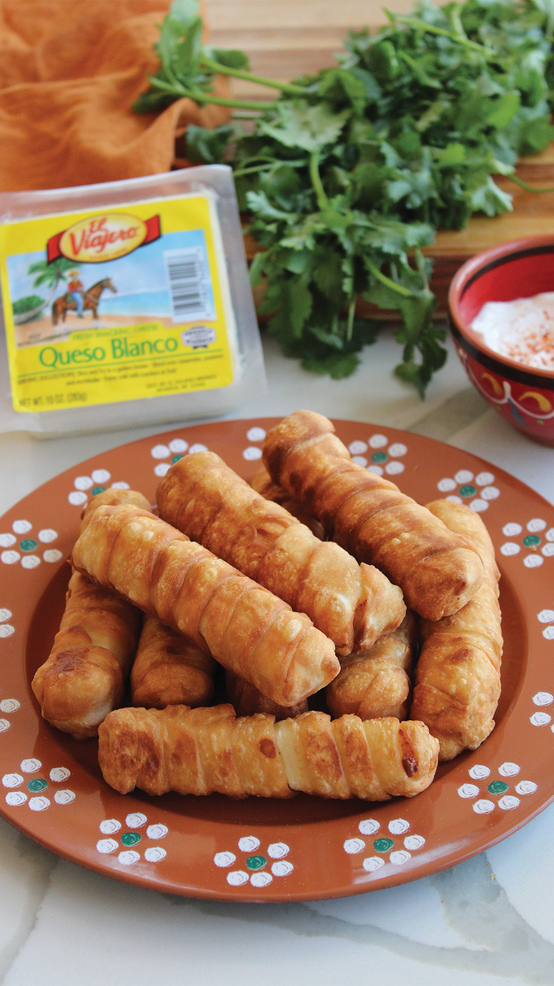 A plate of fried taquitos with a side of shredded cheese, surrounded by fresh cilantro, queso blanco cheese, and a bowl of salsa.