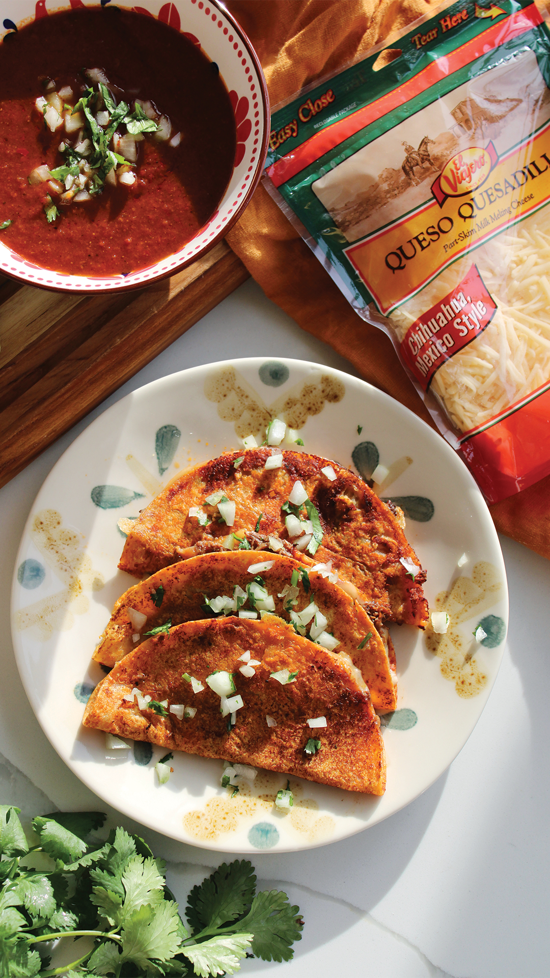 Three Mexican-style tacos on a white plate garnished with chopped onions and cilantro, with a bowl of red sauce and shredded cheese nearby, and a bunch of cilantro at the bottom left corner.