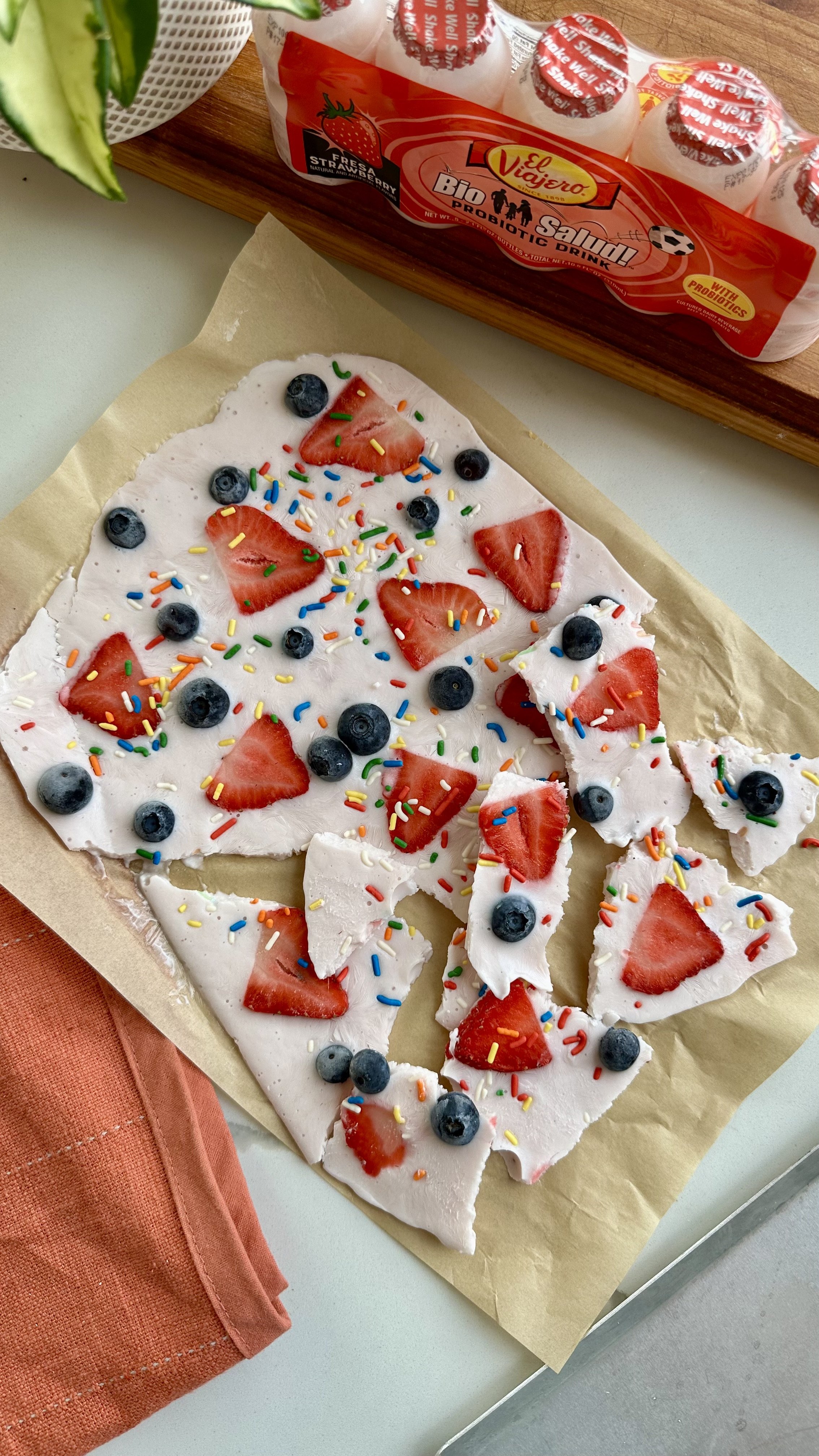 Un broken fruit pizza topped with strawberries, blueberries, colorful sprinkles, on parchment paper. A pack of probiotic drinks and an orange cloth napkin are also visible.