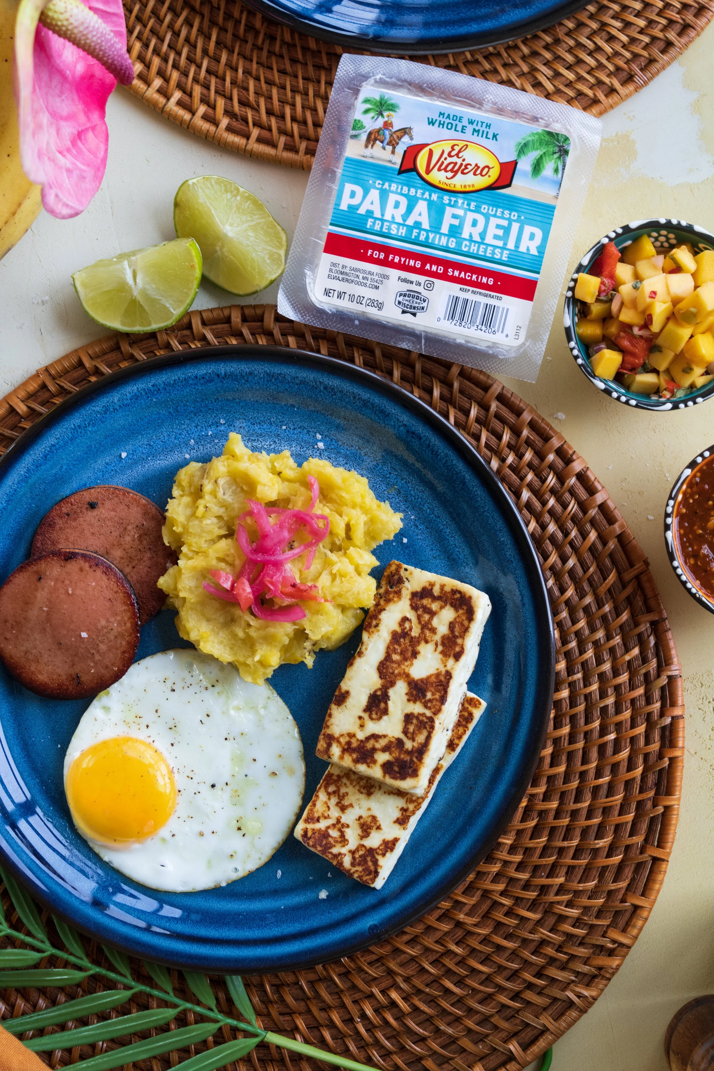 A breakfast plate with a fried egg, two slices of sausage, mashed potatoes with pickled onions, and grilled cheese. There are lime wedges and salsa bowls on the table, along with a package of cheese and some fruit salad.