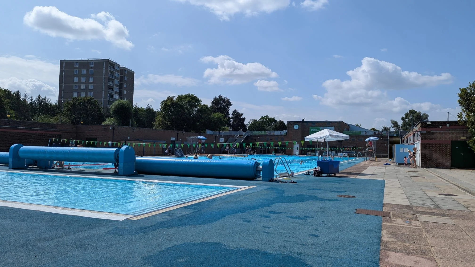 An outdoor public swimming pool under a partly cloudy sky, with pool covers partially rolled up, surrounded by a paved area, umbrellas, and a few people.