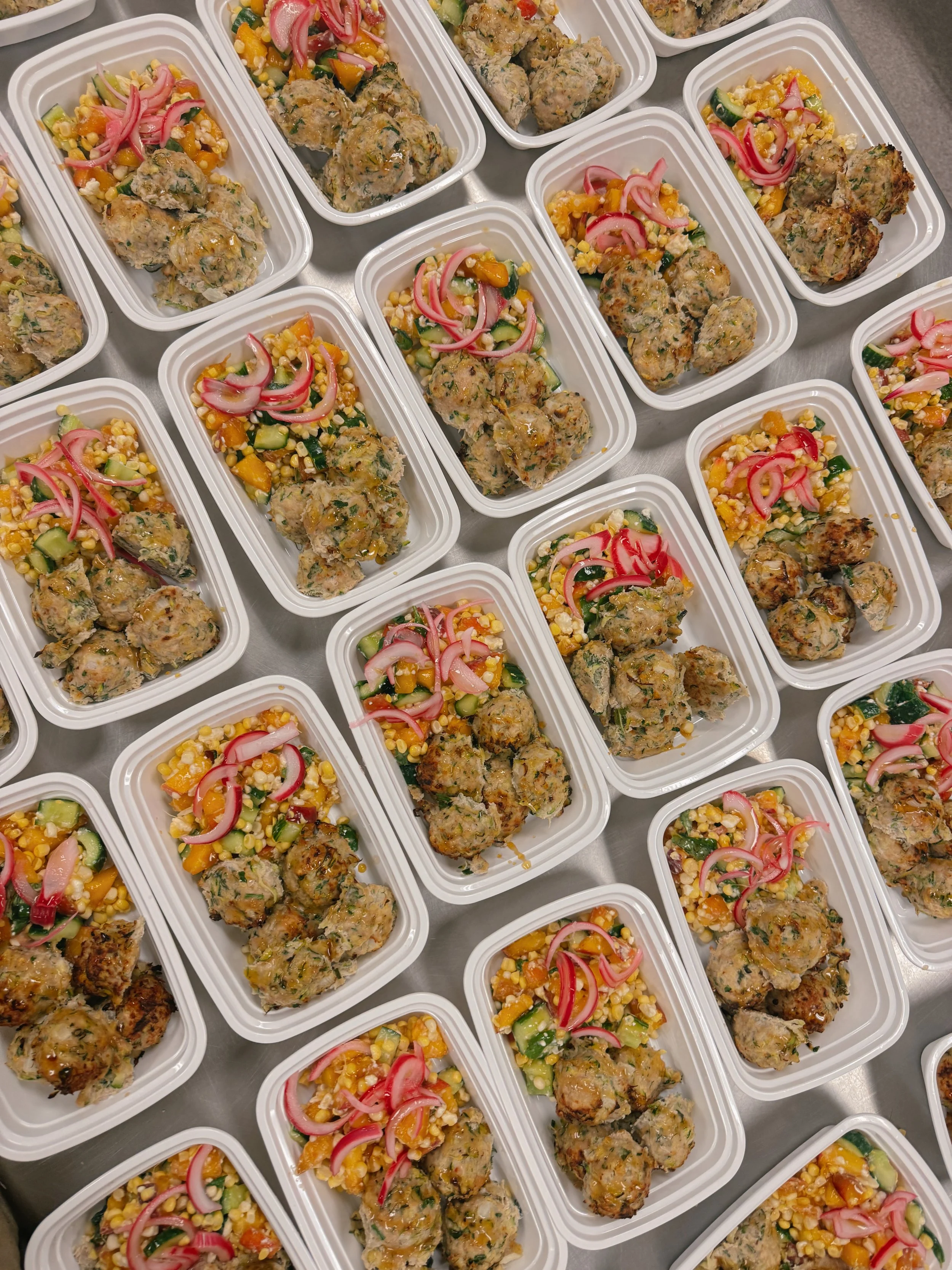 Multiple white food containers on a table filled with stuffed meatballs and colorful vegetable salad garnished with pickled red onions.