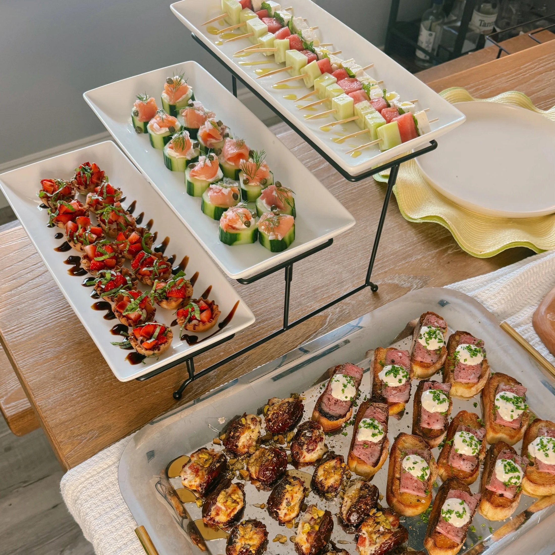 Assorted appetizers displayed on white platters at a buffet, including skewers with melon, watermelon, and cheese, cucumber cups topped with salmon and cream cheese, and various slices of meat and bread with toppings.