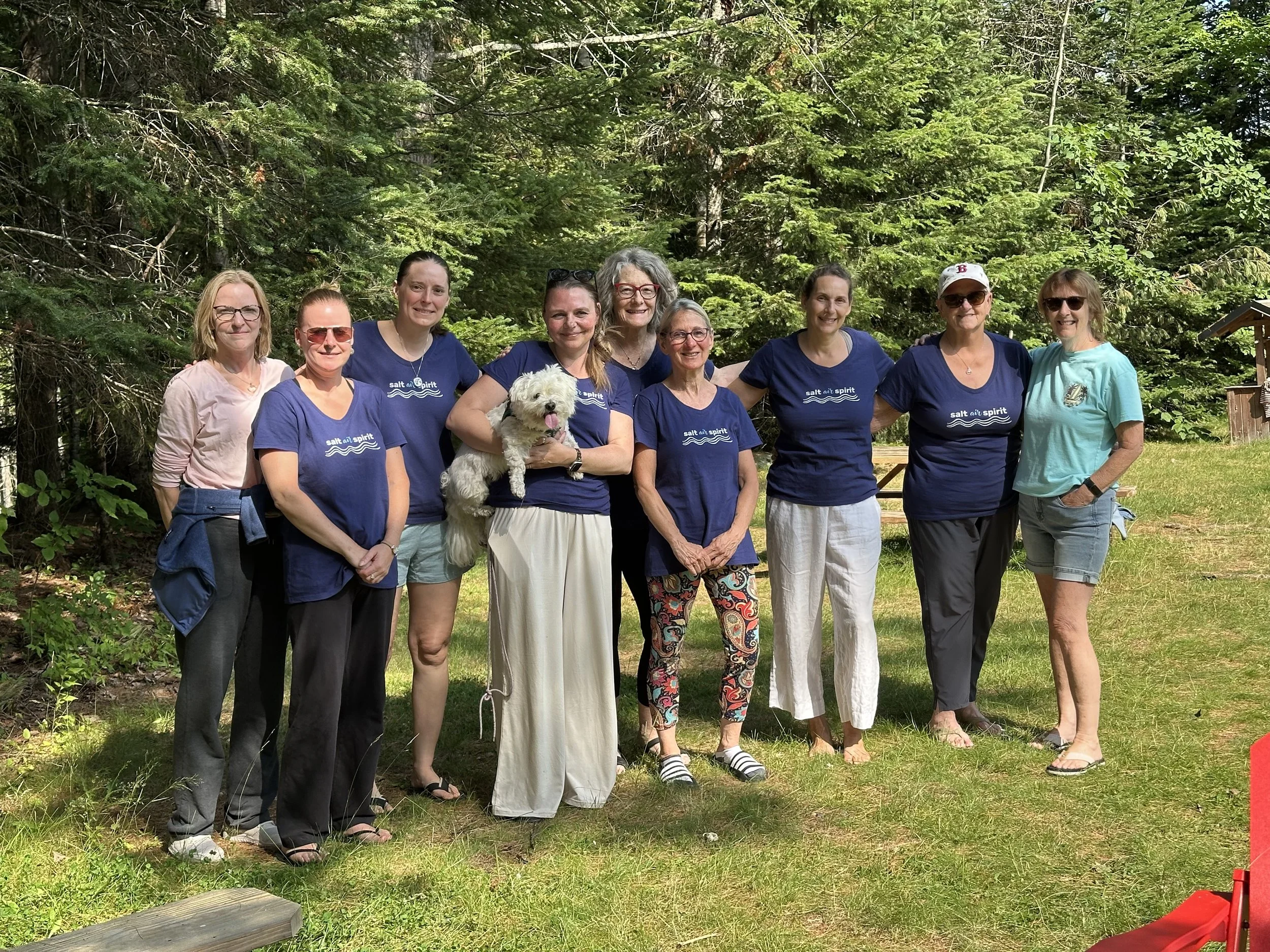Silent retreat Group of eight women with one dog standing outdoors in a forested area with green trees in the background, smiling and posing for a photo.