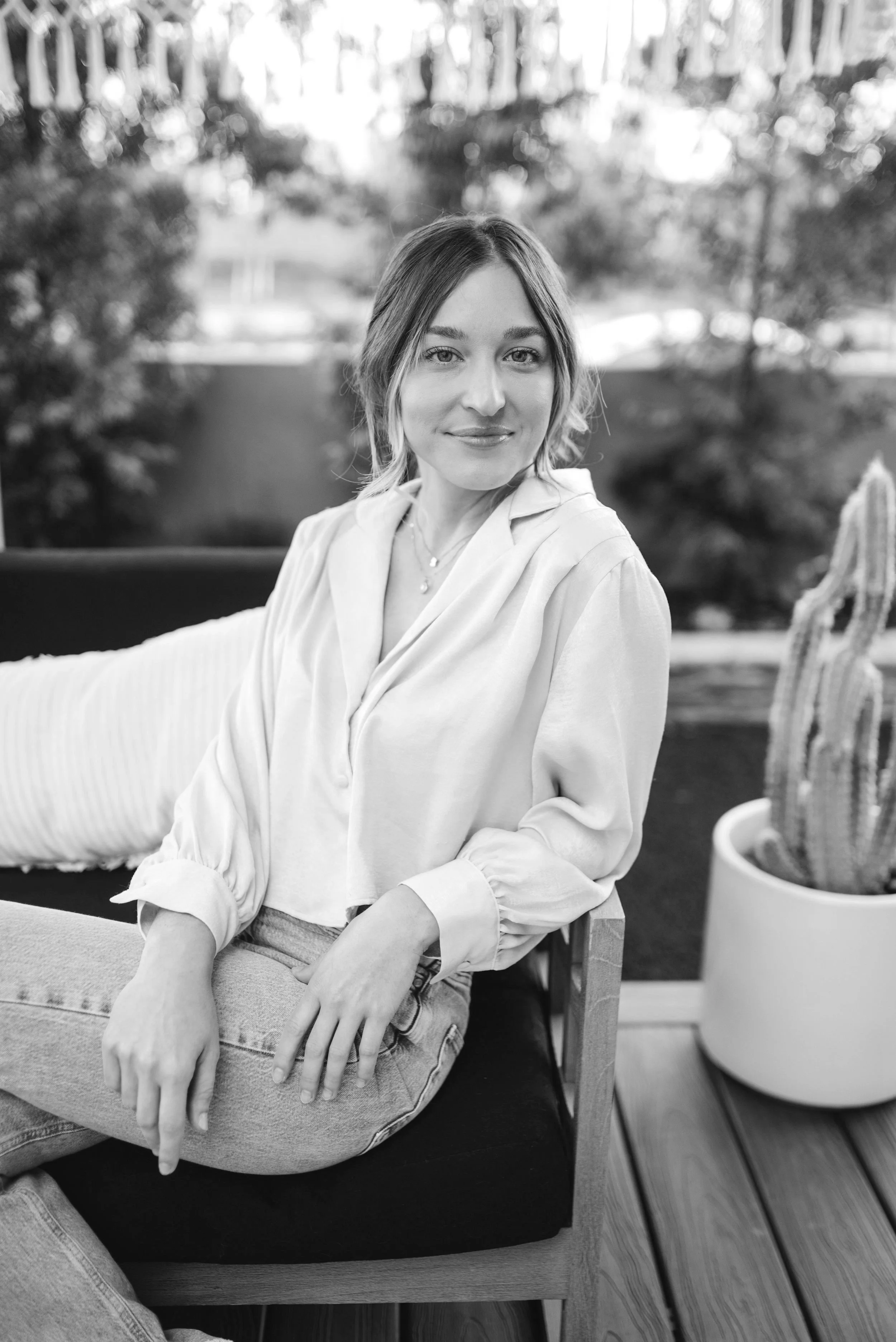 A woman with shoulder-length hair, wearing a light-colored blouse and jeans, sitting on a chair indoors with a smiling expression, next to a potted cactus plant.