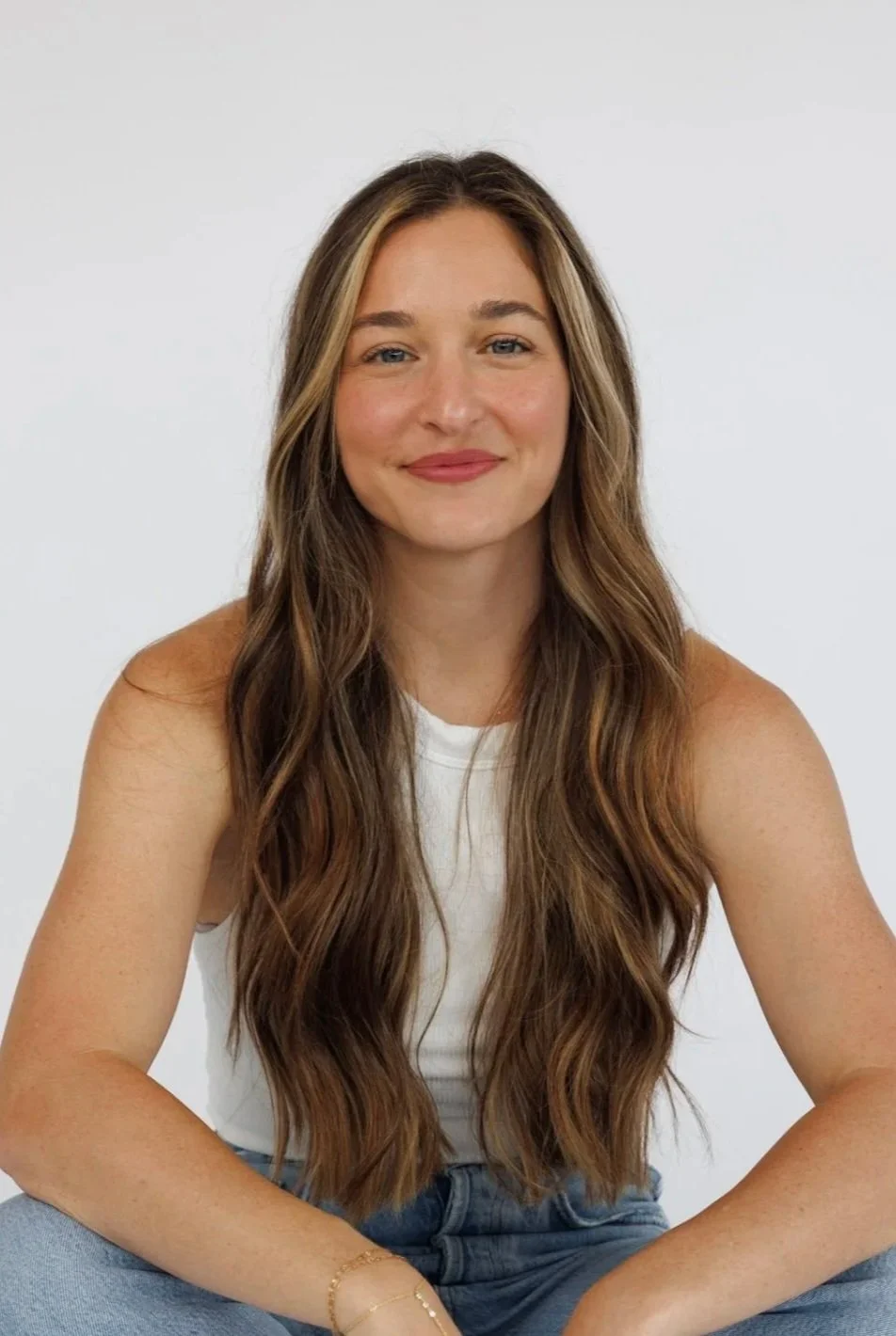 A young woman with long wavy brown hair, smiling, wearing a white sleeveless top and light blue jeans, sitting against a plain white background.