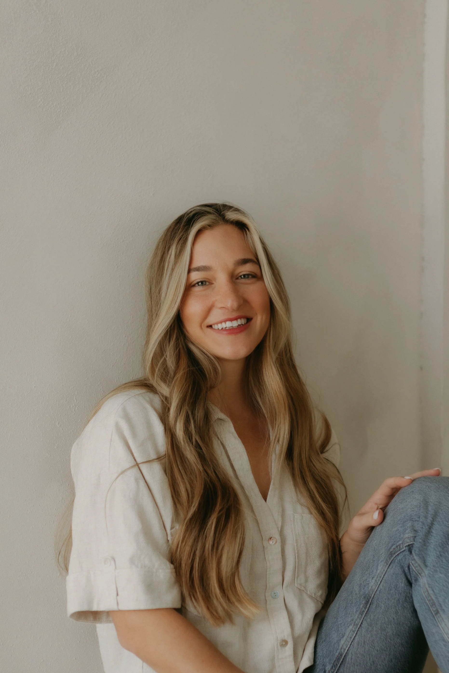 A young woman with long, wavy blonde hair, wearing a beige blouse and smiling, sitting against a plain off-white wall.