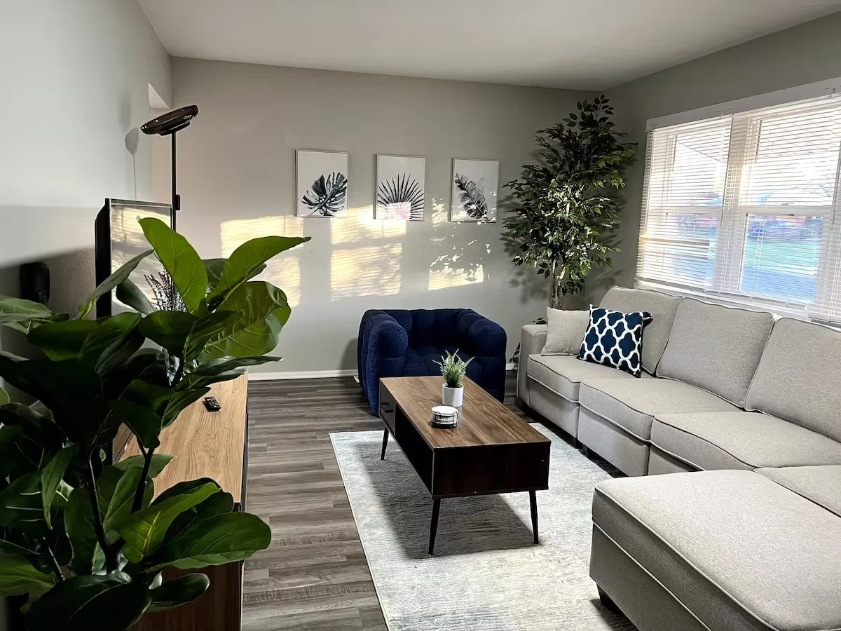 Living room with a beige sectional sofa, blue armchair, wooden coffee table, green plants, wall art with leaf prints, and a window with blinds.
