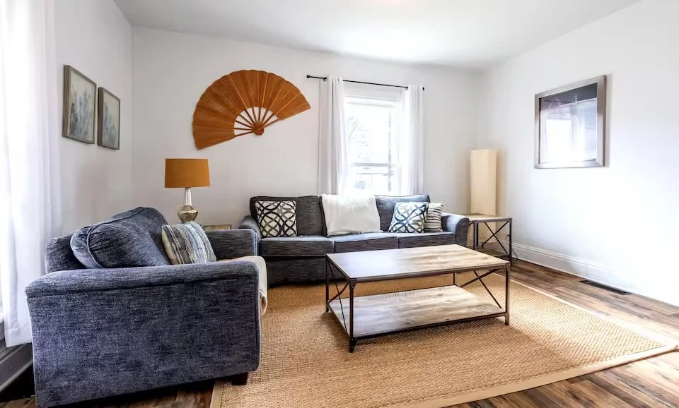 Living room with two dark gray couches, a wooden coffee table, a beige area rug, framed artwork, a tall floor lamp, a table lamp, a window with white curtains, and a wall decoration resembling a fan.