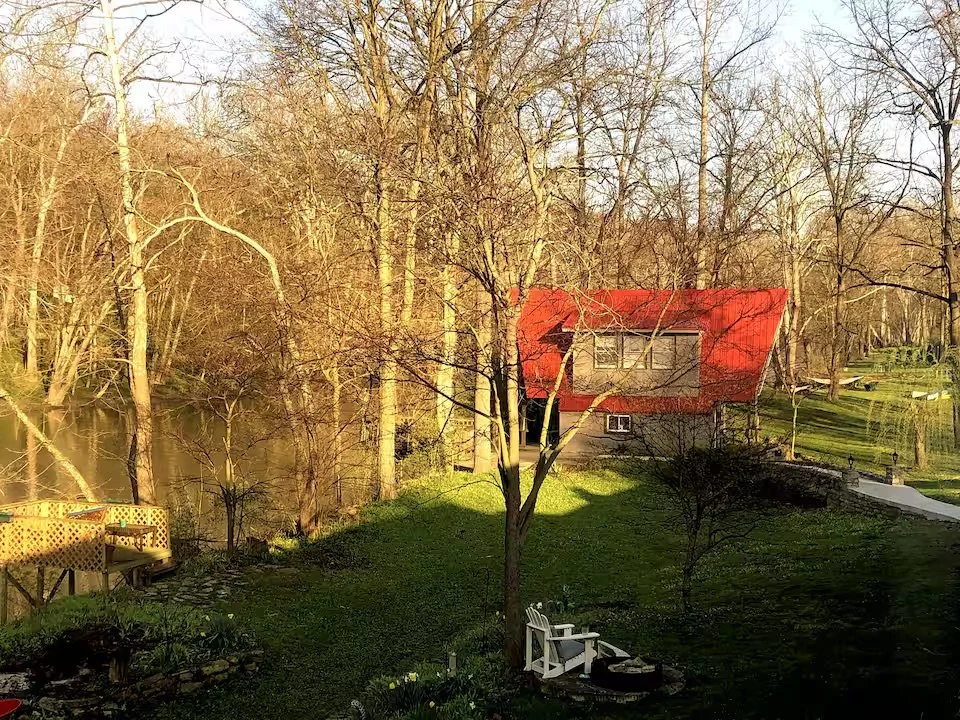 A backyard with trees, grass, a small lake, a white bench, and a house with a red roof in the background.