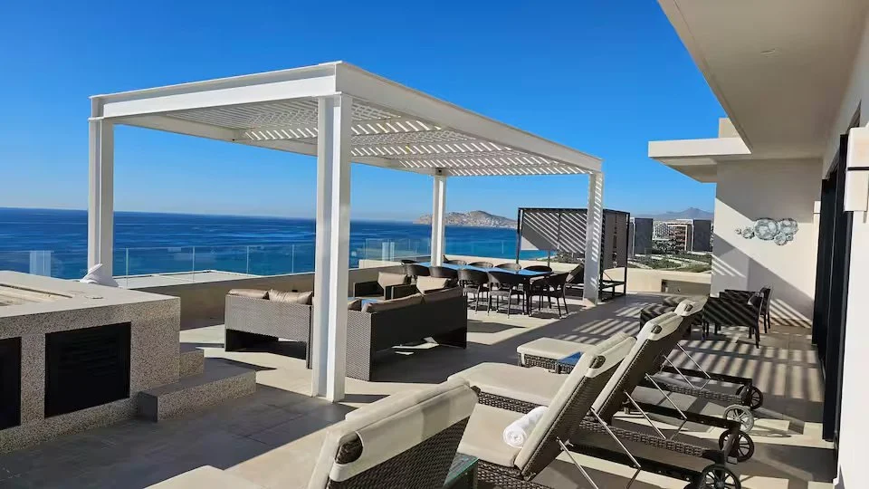 Luxury rooftop patio with lounge chairs, dining table, and seating area overlooking the ocean and city skyline on a sunny day.